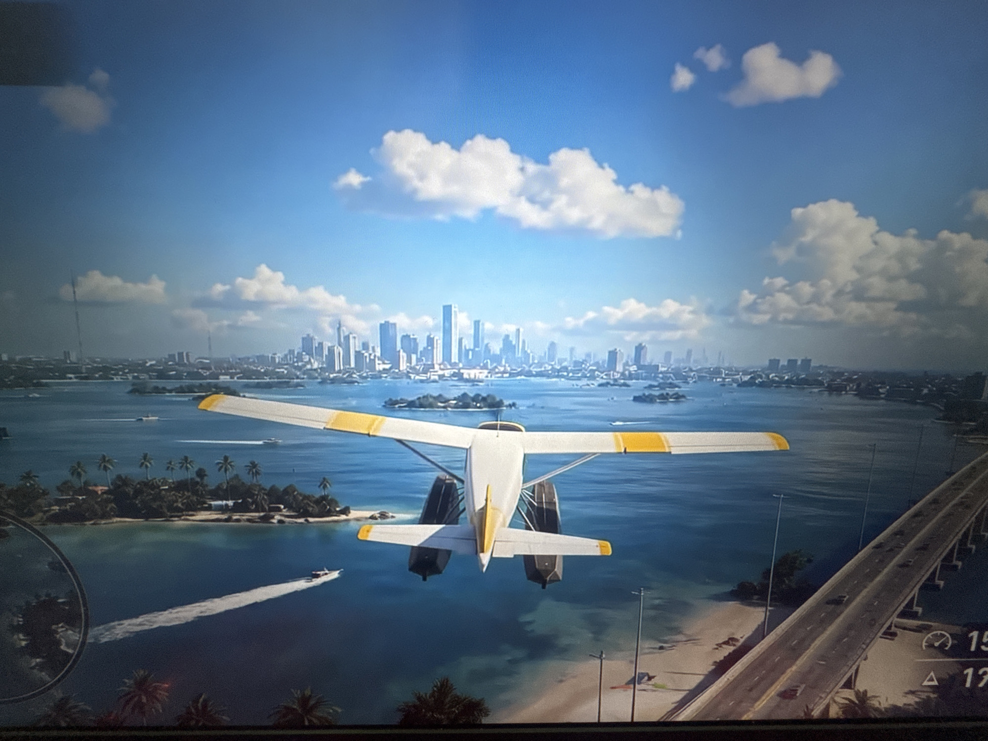 The image depicts a scenic view from an aerial perspective, likely from a video game. In the foreground, a white seaplane with yellow accents on its wings and tail is in flight over a body of water. The plane has two floats attached to its underside. In the background, a city skyline is visible, characterized by numerous skyscrapers. The body of water is dotted with small islands, some of which are covered in palm trees. A long bridge with cars driving on it stretches across the water towards the city. The sky is blue with scattered white clouds. The bottom portion of the image shows a glimpse of a sandy beach with palm trees and some debris. There is also a circular element in the lower left corner, possibly part of a user interface. The image does not show any elements related to Stepnogorsk city administration, Kazakhstan.