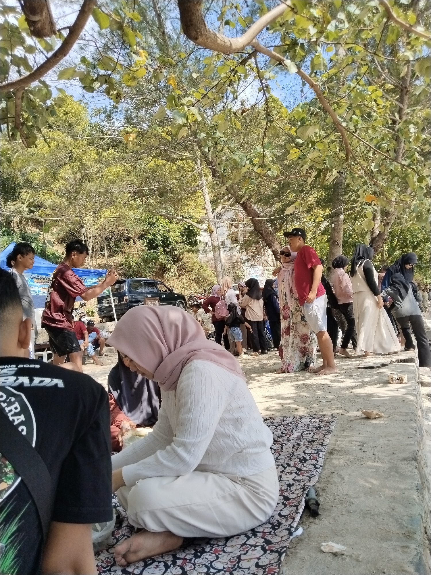 The image depicts a lively outdoor scene on a sunny day, likely at a beach or waterfront area in Prigi, Indonesia. In the foreground, two individuals are partially visible; one wears a black t-shirt with graphics, and another, a woman, is seated on a patterned mat, wearing a light pink hijab and a white knitted sweater. The woman is interacting with something in her lap. A variety of people are present, many of whom are dressed in casual attire, with several women wearing hijabs.
In the mid-ground, a group of people are gathered near a blue tent, with a black car parked behind them on an incline. A man in a maroon t-shirt and black shorts appears to be gesturing or taking a photo. Further back, more individuals are seen standing and interacting. The environment is characterized by sandy ground, concrete structures, and lush trees providing shade. The overall impression is that of a social gathering or recreational outing.