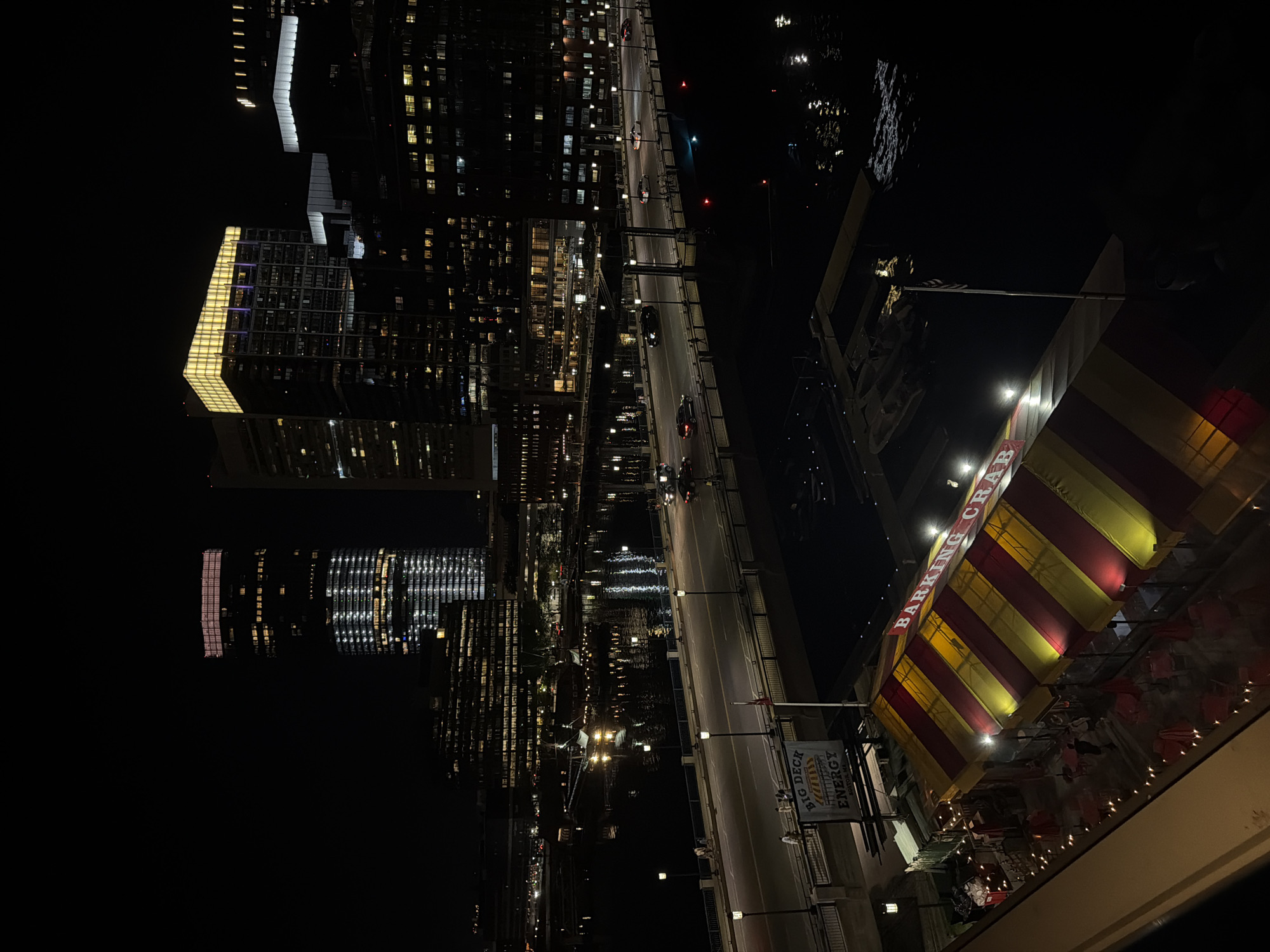At night, the image shows a view from a high elevation overlooking the city of Boston, Massachusetts. The scene includes several tall buildings illuminated with lights, a bridge with moving car traffic, and a building with a striped awning labeled "Barking Crab." A sign for "Big Deck Energy" is also visible near the "Barking Crab" building. The dark water reflects the lights of the city.