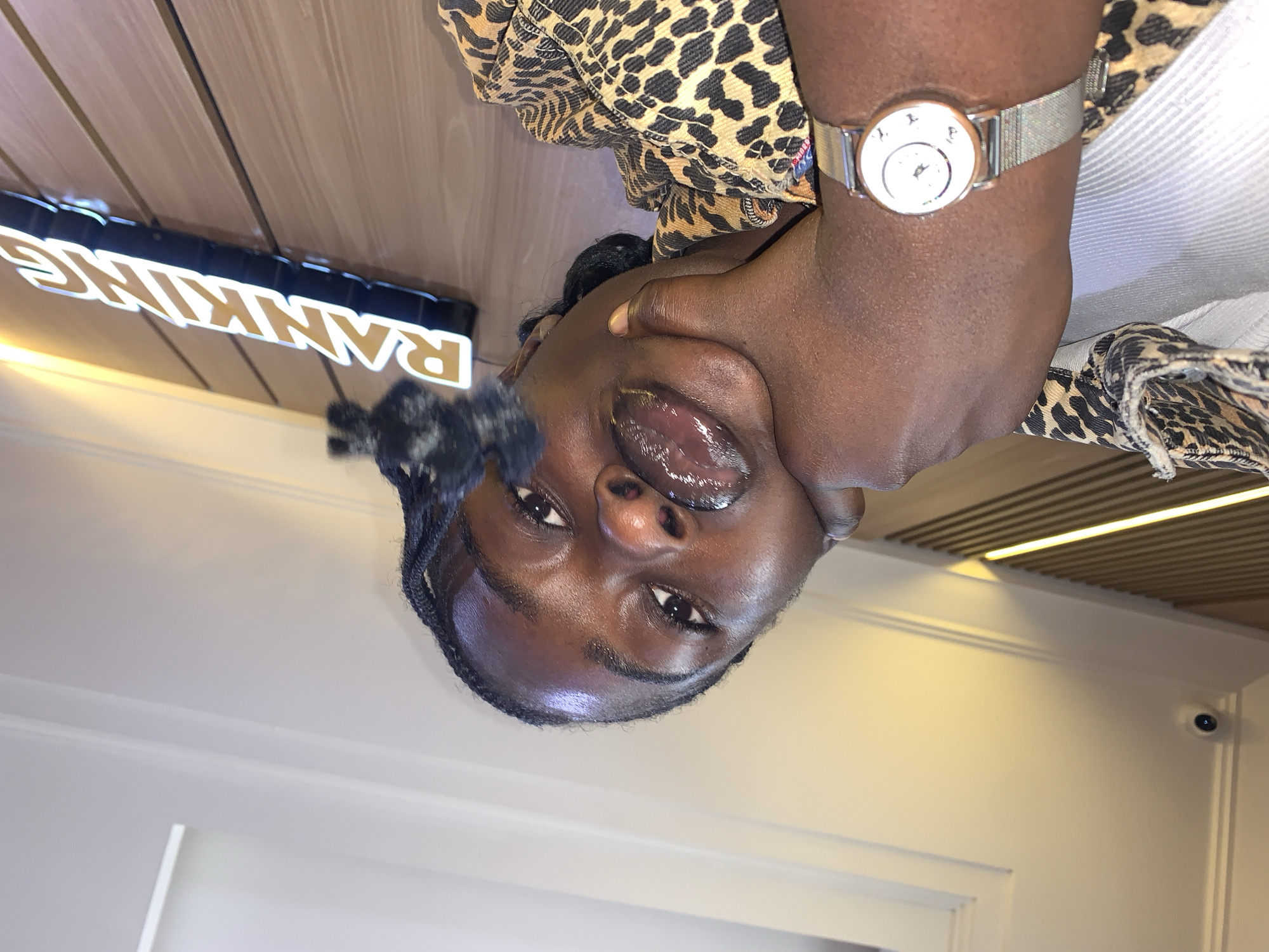 The image shows a close-up of a young Black woman with her face tilted upwards. She is wearing a leopard-print top and a silver watch with a mesh band on her left wrist. Her mouth is slightly open, and she appears to be looking upwards. In the background, there is a sign with illuminated letters that spell out "RANKING". The setting appears to be indoors, with wooden paneling and lighting fixtures visible. There is also a security camera mounted on a wall in the background. The overall tone of the image is intimate and personal, with the subject's gaze drawing the viewer in. The lighting is soft and warm, creating a relaxed atmosphere. The image does not provide clear cues about the time of day or weather, but the indoor setting suggests it could be any time.