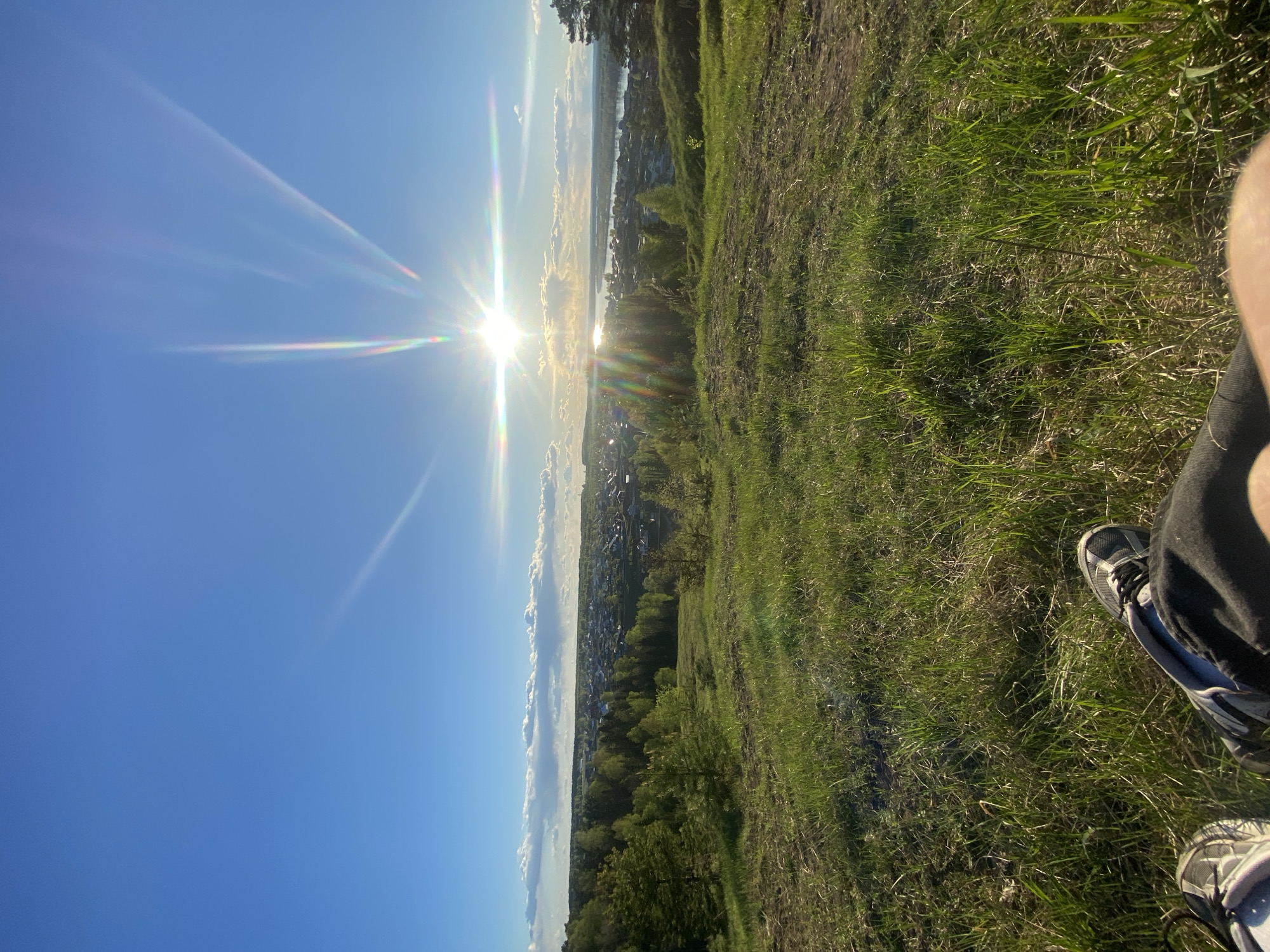 The image presents a daytime scene at a high-angle view, likely from a hilltop, overlooking a landscape. A bright sun with distinct rays dominates the sky, appearing to set or rise over a distant area that includes a possible body of water and a town or city.
The landscape in the midground includes trees and what appears to be a long stretch of buildings and houses, indicative of an urban area. In the foreground, there is grassy terrain, suggesting a natural or semi-natural environment on the hilltop. The presence of shoes and part of a leg in the lower right of the image suggests someone is sitting or resting on the hilltop, taking in the view.
The details specific to Ольховка, Russia, are challenging to confirm from the visual information alone. The city's exact features or landmarks are not discernible without additional geographic clues or contextual data.