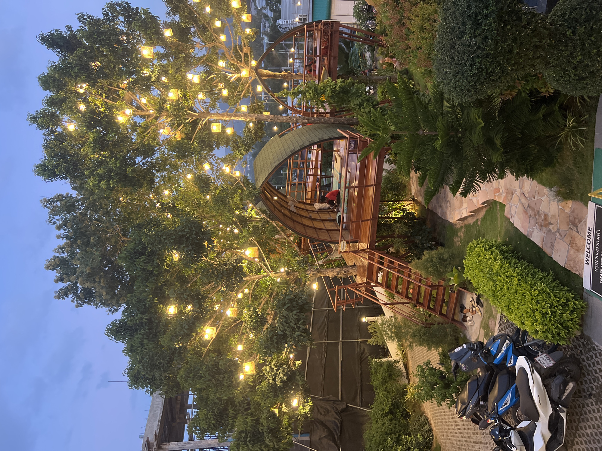 The image depicts an outdoor seating area at dusk, likely a restaurant or cafe, nestled within a tropical setting on the island of Ko Samui, Thailand. The scene is characterized by two elevated, dome-shaped wooden structures with thatched roofs, reminiscent of treehouses, built around large trees. These structures are adorned with numerous yellow string lights that illuminate the branches and create a warm ambiance.
One of the dome structures, on the left, has a wooden walkway and stairs leading up to it. Inside, a person wearing a red shirt is seated on a floor cushion, facing away from the camera. The other dome structure, on the right, is similarly elevated and also appears to be a seating area, with people visible inside at tables.
The ground level features a paved pathway winding through lush greenery, including hedges and ornamental trees. Several motorcycles are parked on a patterned pavement area in the foreground, with a white and blue scooter prominently displayed on the left. Near the stairs of the left dome, a collection of footwear is arranged on the ground.
In the background, hints of urban development can be seen, including unfinished concrete structures, suggesting the location is situated within or near a developed area. The sky is a muted blue, indicating the transition from day to night, with distant hills visible on the horizon. The overall impression is one of a serene and inviting evening dining experience in a tropical environment.
