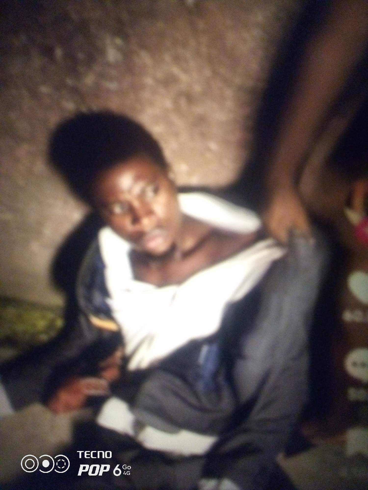 The image, taken at night in Howayi, Nigeria, shows a person in the foreground with their shirt pulled up, exposing their chest. Their head is turned slightly to the side, and their eyes are open. Another person's arm is visible in the upper right corner, reaching towards the individual in the foreground. The background is blurred and appears to be an outdoor setting with a rough, textured surface. A "TECNO POP 6 4G" watermark is present in the lower left corner.
