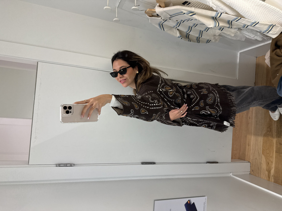 A woman in sunglasses poses for a mirror selfie. She wears a brown patterned jacket, grey jeans, and white sneakers. Her left hand holds a cell phone. The background includes clothing racks with white and blue garments, a white door, and a wooden floor. A poster is visible on the wall. The image appears to be taken inside a store or dressing room.