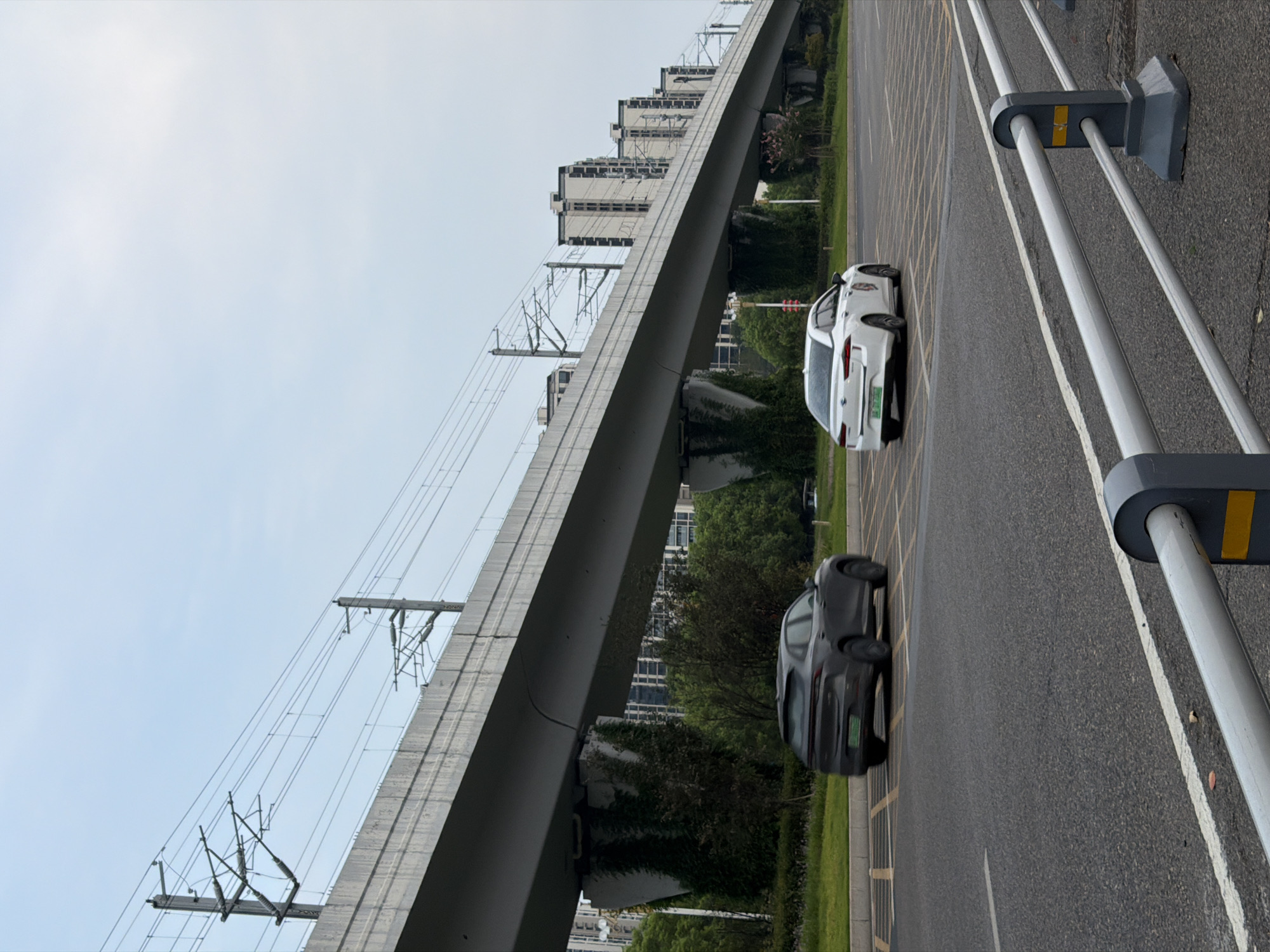 The image shows a street scene in Jiangjin, China, under an elevated railway or highway structure. Two cars are visible on the road: a dark-colored SUV in the left lane and a white sedan in the right lane. The elevated structure, supported by concrete pillars partially covered in ivy, extends into the background, where a series of residential buildings are present. Overhead, power lines for the railway or highway are suspended between poles. The sky is overcast. The foreground features a metal guardrail along the edge of the road.