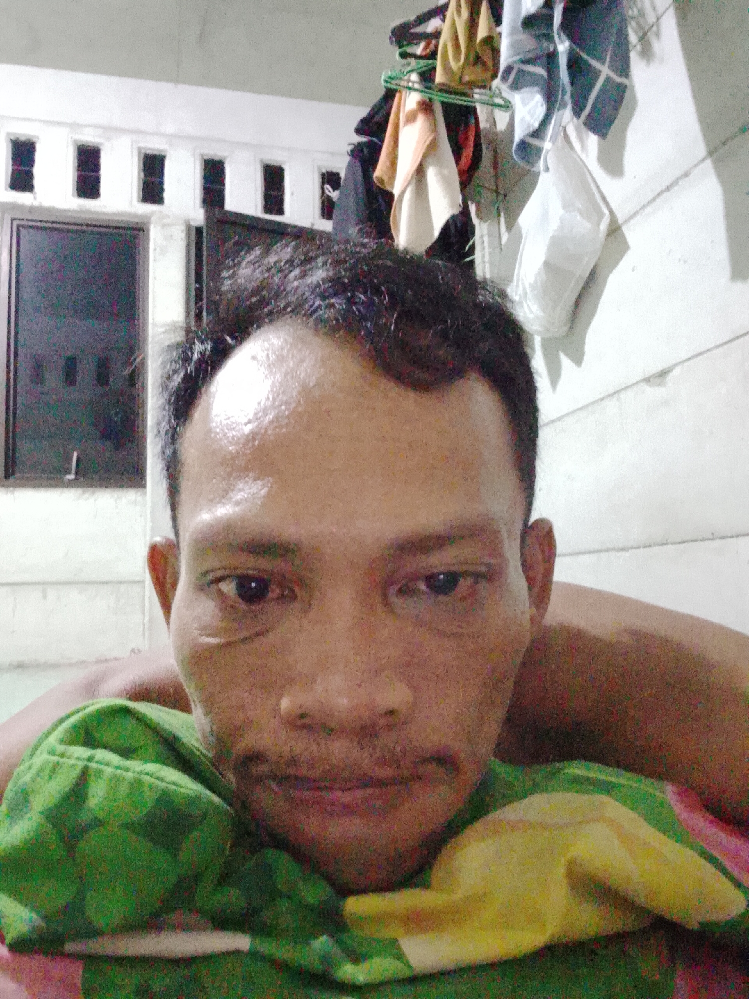 A close-up, indoor selfie captures a man lying down with his head near the bottom of the frame. The man has dark hair, a broad face, and appears to have a slight sheen on his forehead. He is wearing a green shirt with yellow patterns. Behind the man, a white wall is visible with a window in the upper-left corner containing several smaller panes. Hanging on a rack against the wall are articles of clothing and a white face mask. The location is potentially in the city of Bukit Ketok, Indonesia.