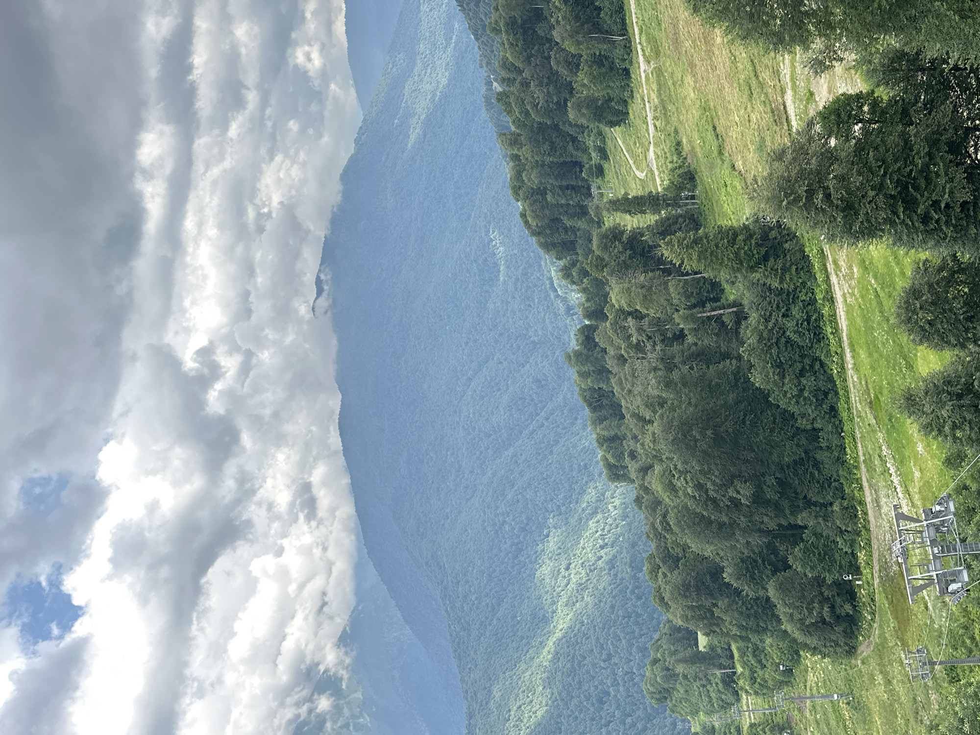 The image shows a panoramic view of a mountainous landscape in Estosadok, Russia. In the foreground, a grassy slope is dotted with trees, and a ski lift structure is visible. A dirt path winds its way up the slope, disappearing into the dense green forest that covers the lower and middle sections of the mountains. The middle ground is dominated by rolling hills covered in lush green trees, with some areas receiving dappled sunlight. In the background, a series of larger, hazy blue mountains recede into the distance, their peaks partially obscured by clouds. The sky is overcast with a mix of dark and light gray clouds, with a small patch of blue sky visible in the upper left quadrant. The overall impression is a serene, natural environment, likely a mountain resort during the summer or early autumn.