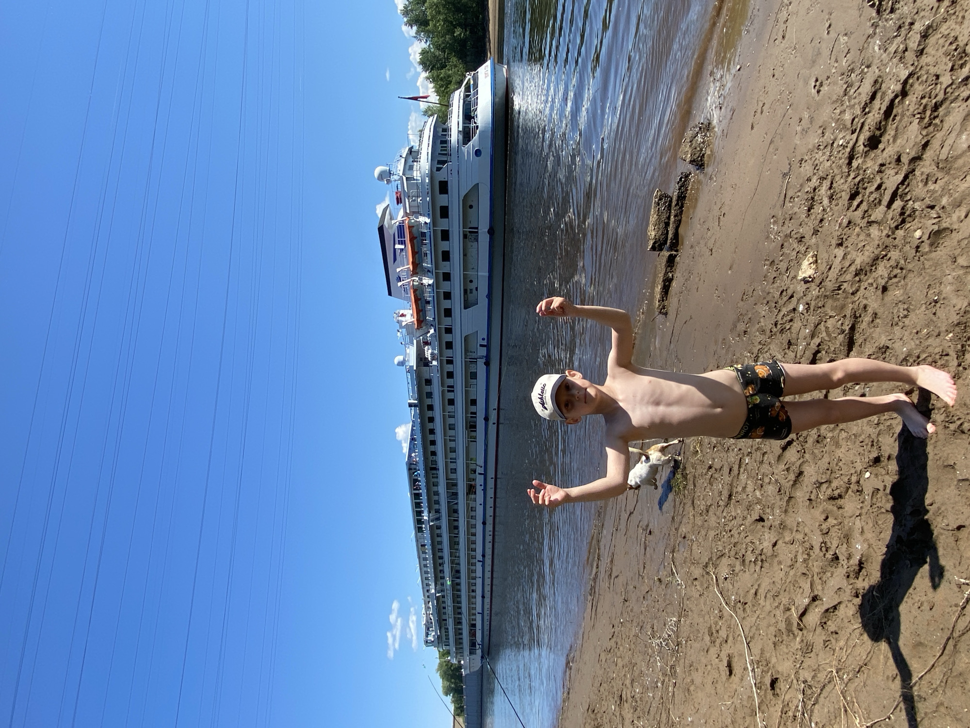 A young boy stands on a sandy riverbank, facing the camera with a slightly raised chin. He is shirtless and wearing patterned swim trunks. His arms are bent upwards, with his hands open. To his left, a large white cruise ship is docked in the river. The ship has multiple decks and several lifeboats are visible along its side. Behind the ship, a line of trees forms the opposite bank. The sky is a clear, bright blue with a few wispy clouds. Power lines are visible against the sky. The sun is shining brightly, casting shadows on the sand, suggesting it is daytime, likely midday. The overall scene depicts a recreational moment by the river, possibly during a vacation or day trip. The boy's pose could indicate a playful mood or a gesture of showing off. There is no visible text in the image.