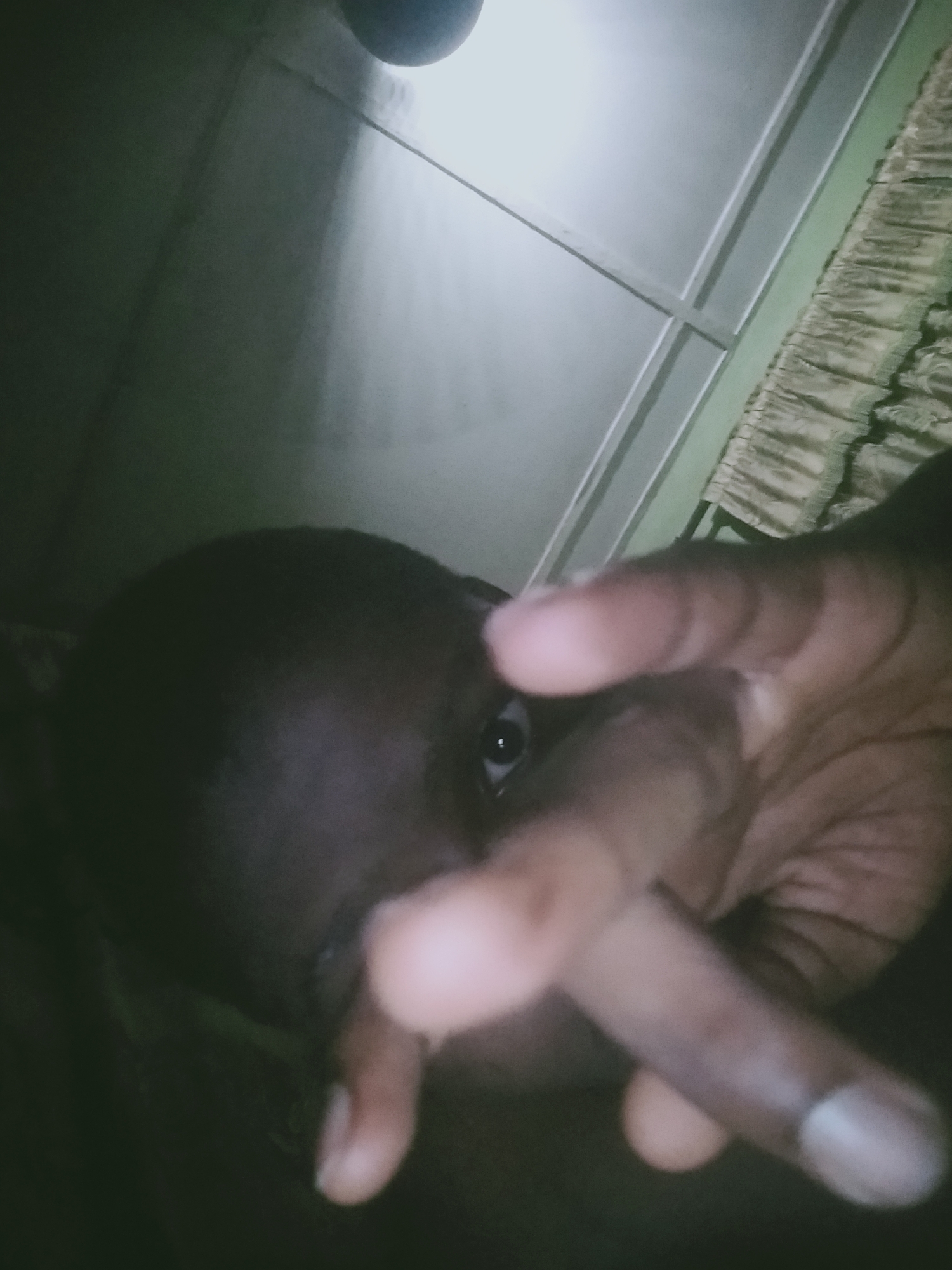 A close-up, low-angle photograph taken indoors. A person with dark skin is in the foreground. Their face is partially obscured by their hand. Only one eye is visible. The hand is brown and the fingers are extended, with the index and middle fingers forming a "V" shape. The background shows a white ceiling with visible beams or panels. Along the top right, a curtain or draped fabric with a textured pattern is visible. The lighting in the room is dim.