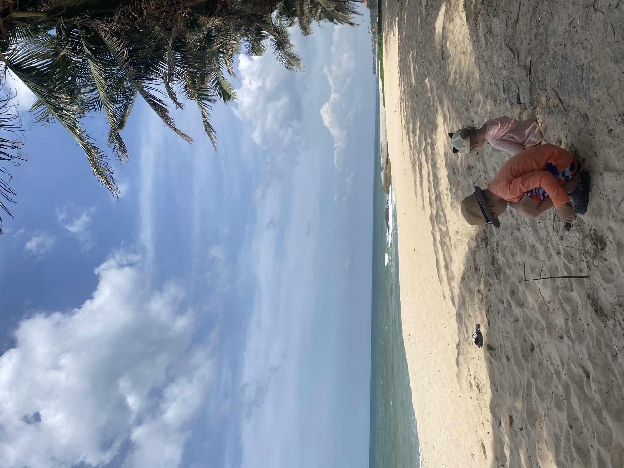 Two children are on a sandy beach in Phu Thuy Ward, Vietnam. The child in the foreground, wearing an orange long-sleeved shirt, patterned shorts, a straw hat, and dark shoes, is crouched down and appears to be playing in the sand. Behind them, a smaller child, wearing a pink hooded garment and a cap with cat ears, is also seated in the sand, facing away from the camera. The beach has fine, light-colored sand and is bordered by the ocean, which has a greenish-blue hue. Palm trees with prominent fronds are visible on the right side of the image, casting shadows on the sand. The sky is partly cloudy with large white cumulus clouds against a blue backdrop. In the distance, the coastline is lined with vegetation and some structures. A single black and white striped sandal lies on the sand near the left edge of the frame. The overall scene depicts a relaxed day at the beach.