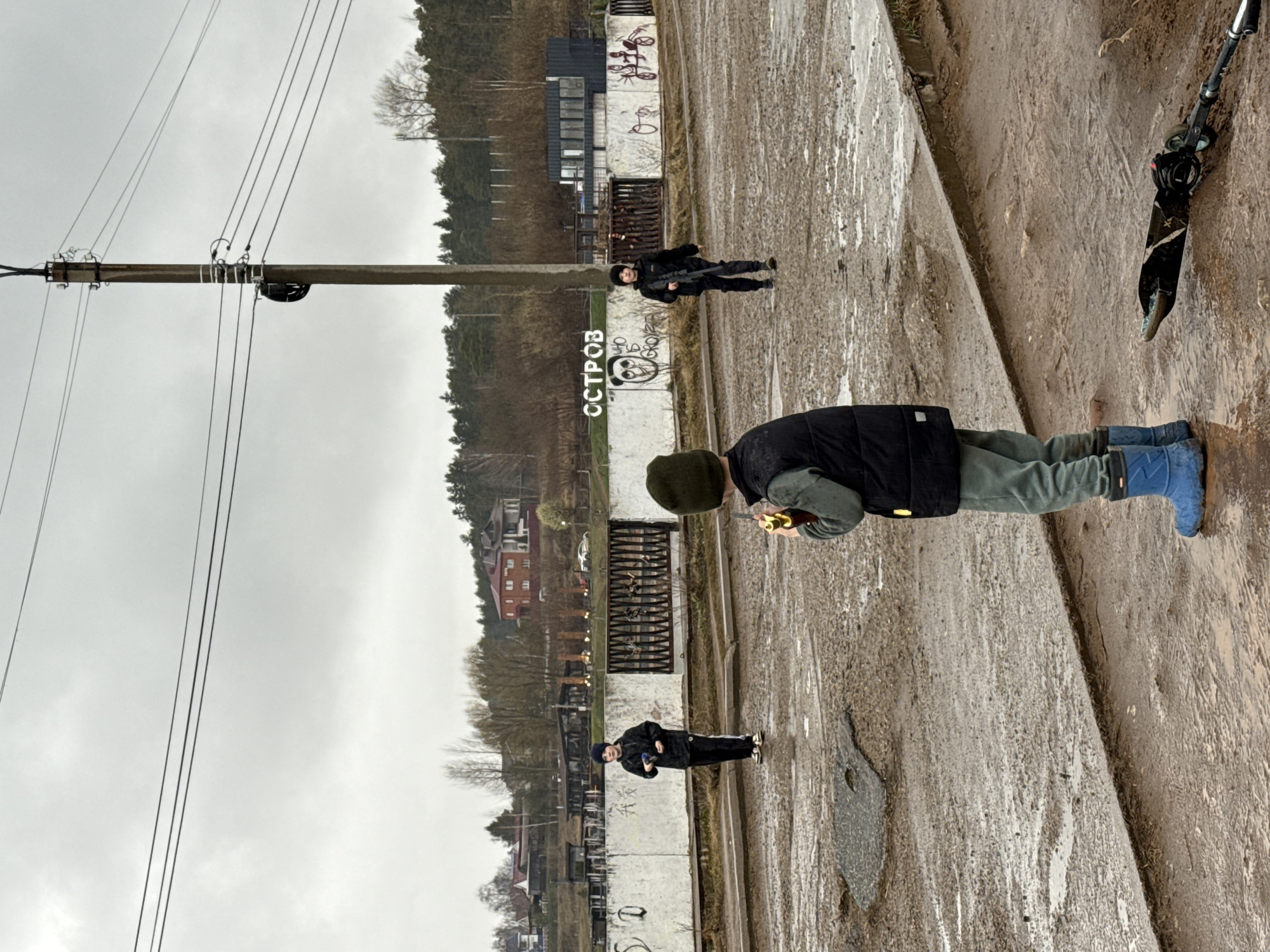 The image shows an overcast day in a location within Votkinsk, Russia. A young boy wearing a green beanie, a black vest, olive-green pants, and blue boots stands on a concrete path, holding an object near his mouth. Two other people, dressed in dark clothing, stand further away near a concrete structure that has 'ОСТРОВ' (Island) written on it in Cyrillic letters. There is also graffiti on the concrete structure. A scooter is leaning against the concrete path. A power pole with numerous power lines stretches into the gray sky. In the background, there are trees and buildings, typical of a Russian town. The ground is wet, suggesting recent rain.