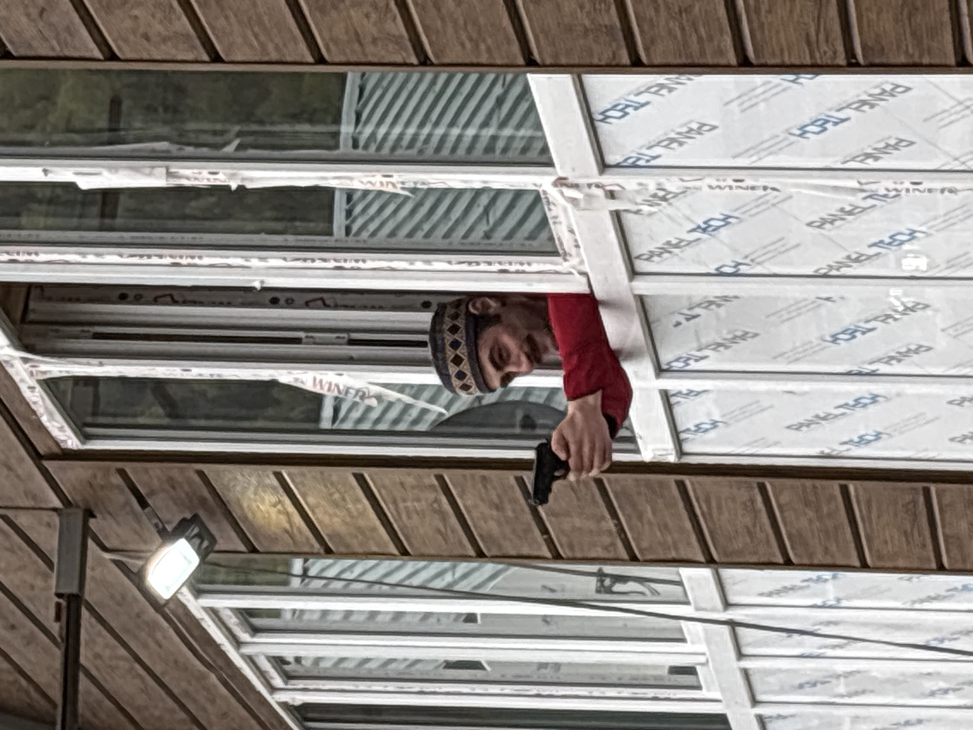 The image shows a man holding a gun while looking through a window. The man is wearing a red shirt and a patterned cap. The window is made up of multiple panes, some of which have a protective film with "PANEL TECH" printed on them. The location is possibly Kveda Chkhutuneti, Georgia, but this is inferred from metadata. The environment appears to be an interior with a roof structure. A light fixture is visible in the lower-left corner.