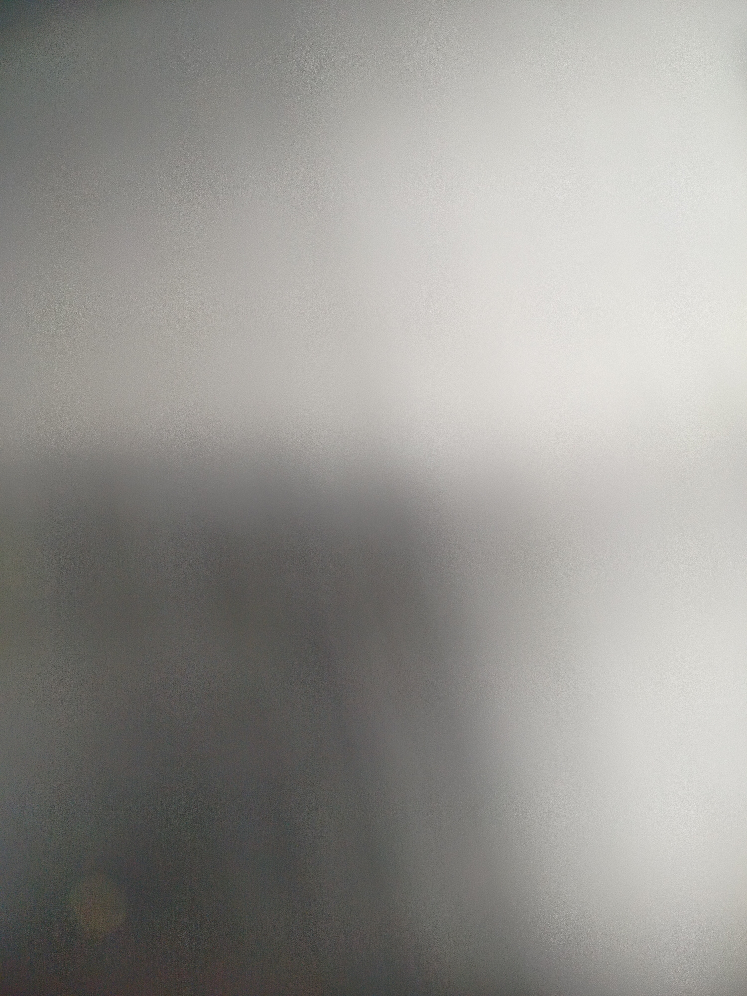 The image shows a blurred and out-of-focus surface, transitioning from a darker gray in the lower left to a lighter gray in the upper right. The texture suggests a matte or brushed material, possibly metal. The lack of distinct features and sharpness makes it impossible to determine the location or identify specific objects or subjects.