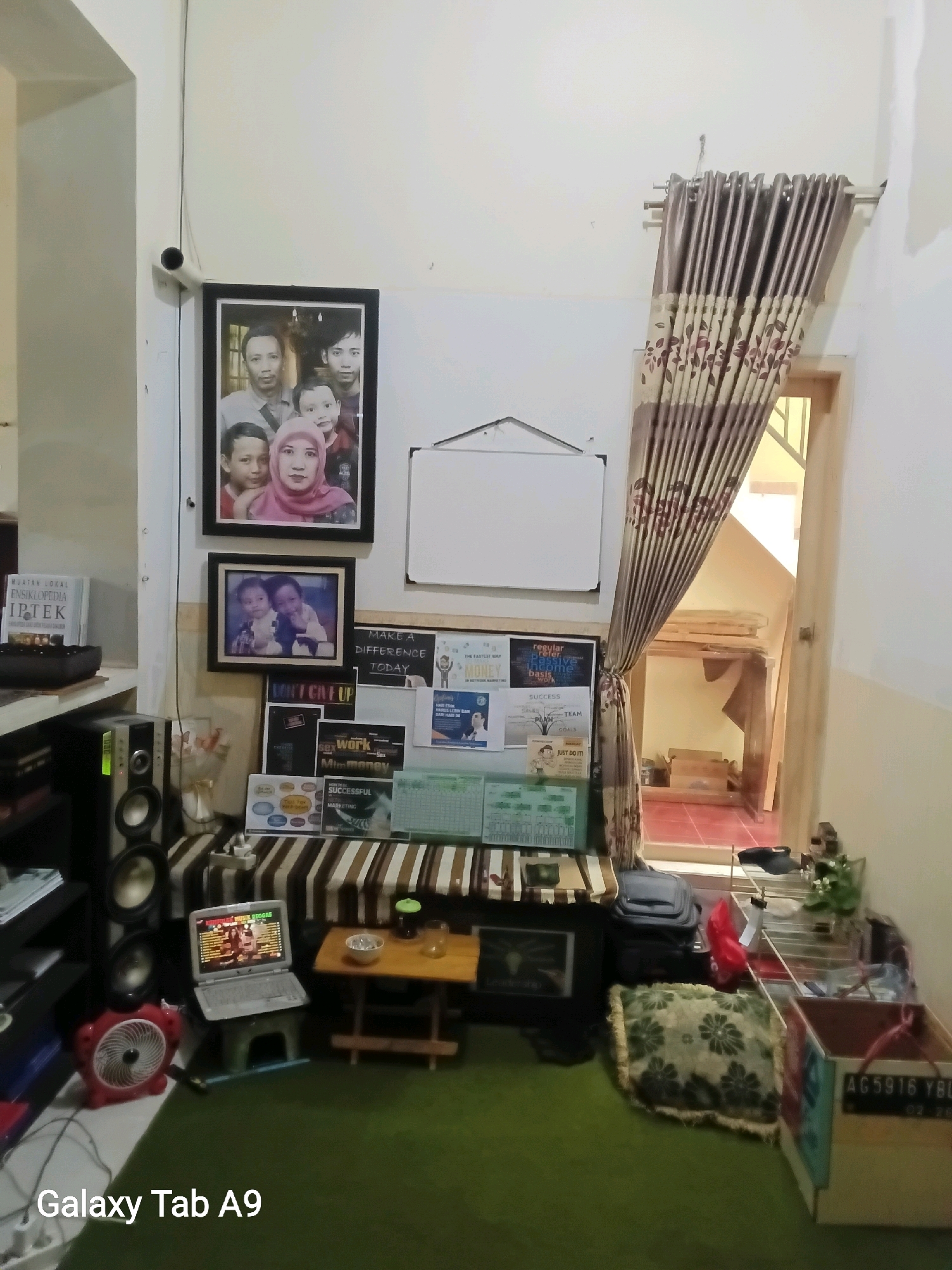 The image depicts an indoor scene, likely a living room or study area, with a carpeted floor and white walls. Several items are visible, including a framed family photograph on the wall, a laptop on a stool, a small wooden table with a drink and a bowl, a fan, speakers, and various motivational posters and books. A window with patterned curtains is also present, revealing a glimpse of the outdoors, which appears to be a staircase leading upwards. The scene is cluttered but organized, suggesting a lived-in space. No specific location in Trenggalek, Indonesia is identifiable from the image itself. The overall impression is of a functional and personalized workspace or relaxation area.