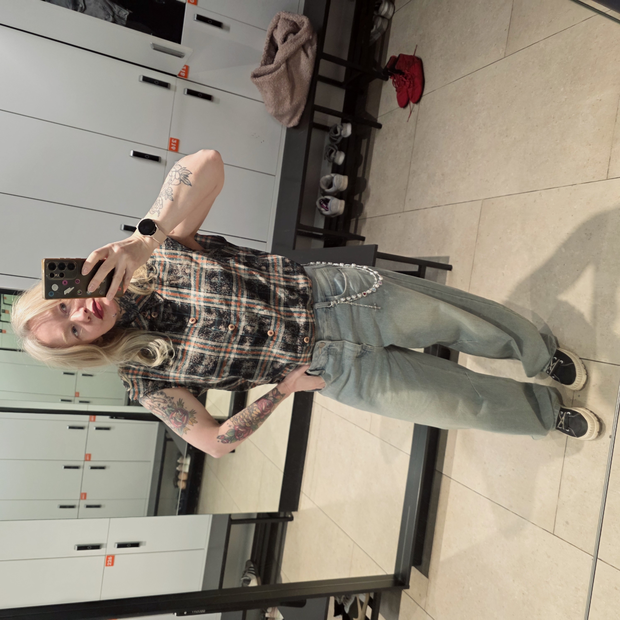 A person with long blonde hair is taking a selfie in a locker room. The person is wearing a plaid shirt, jeans with a chain, and black and white shoes. The person has tattoos on both arms. The locker room has white lockers with orange numbers on them. There is a black bench with shoes on it. There are red shoes on the floor. The flooring is tiled. There is a reflection of the person in the mirror. The location is not apparent from the image.