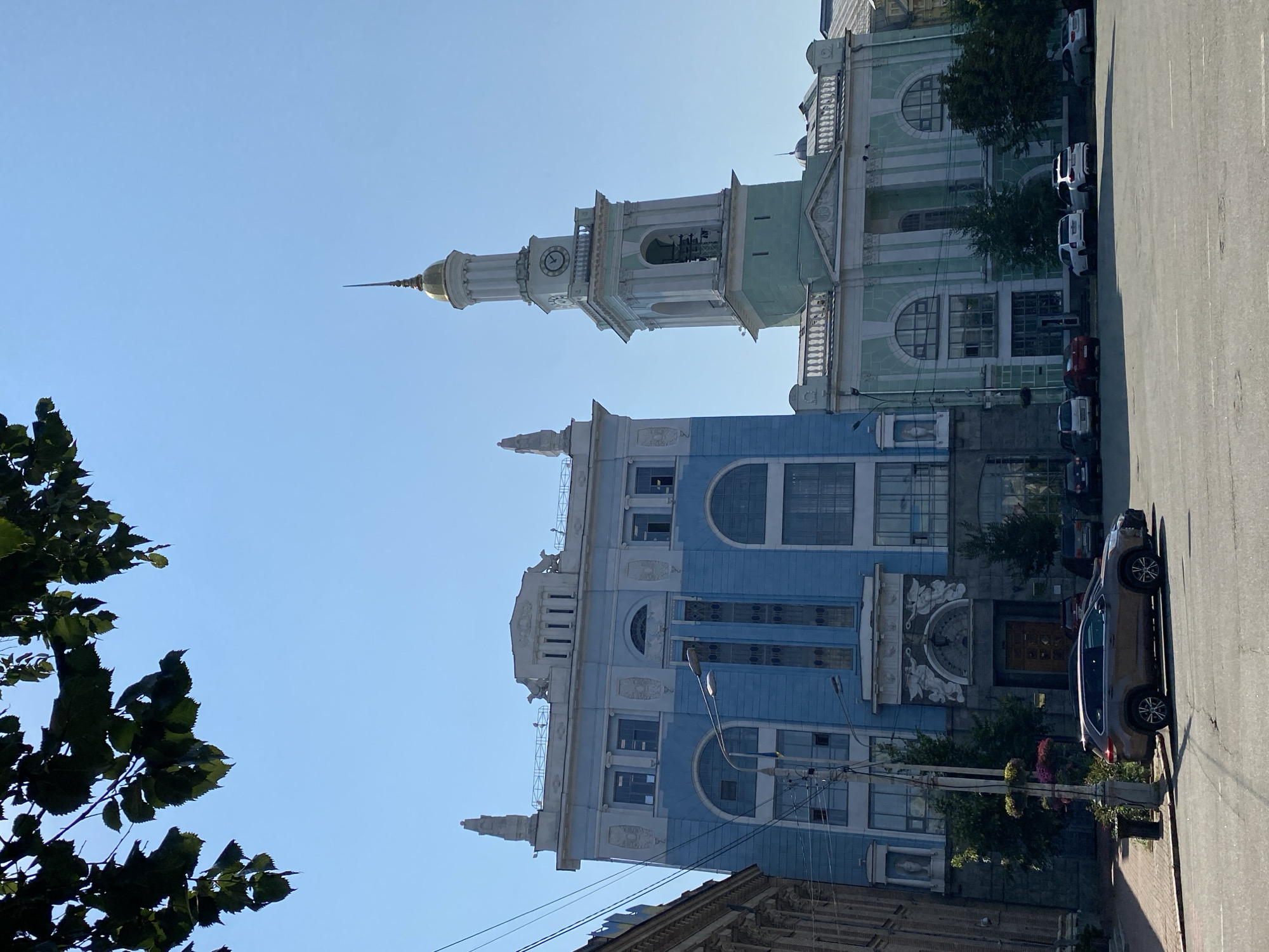 In an urban setting under a clear blue sky, two prominent buildings dominate the view. The building on the left is a light blue, multi-story structure with a distinctive architectural style featuring arched windows and decorative elements. Adjacent to it, to the right, is a pale green building with a tall, slender clock tower topped with a golden dome. The clock face on the tower shows the time as approximately 12:55. Below the tower, there are bells visible within an open structure. The street in front of the buildings is paved and shows some wear. Several cars are parked along the sidewalk and in the street, including a dark grey SUV in the foreground. Green trees and foliage are visible along the sidewalk and around the buildings, adding a touch of nature to the urban landscape. Overhead, tree branches with green leaves partially obscure the sky in the upper left corner of the image. Power lines are also visible, contributing to the city environment.