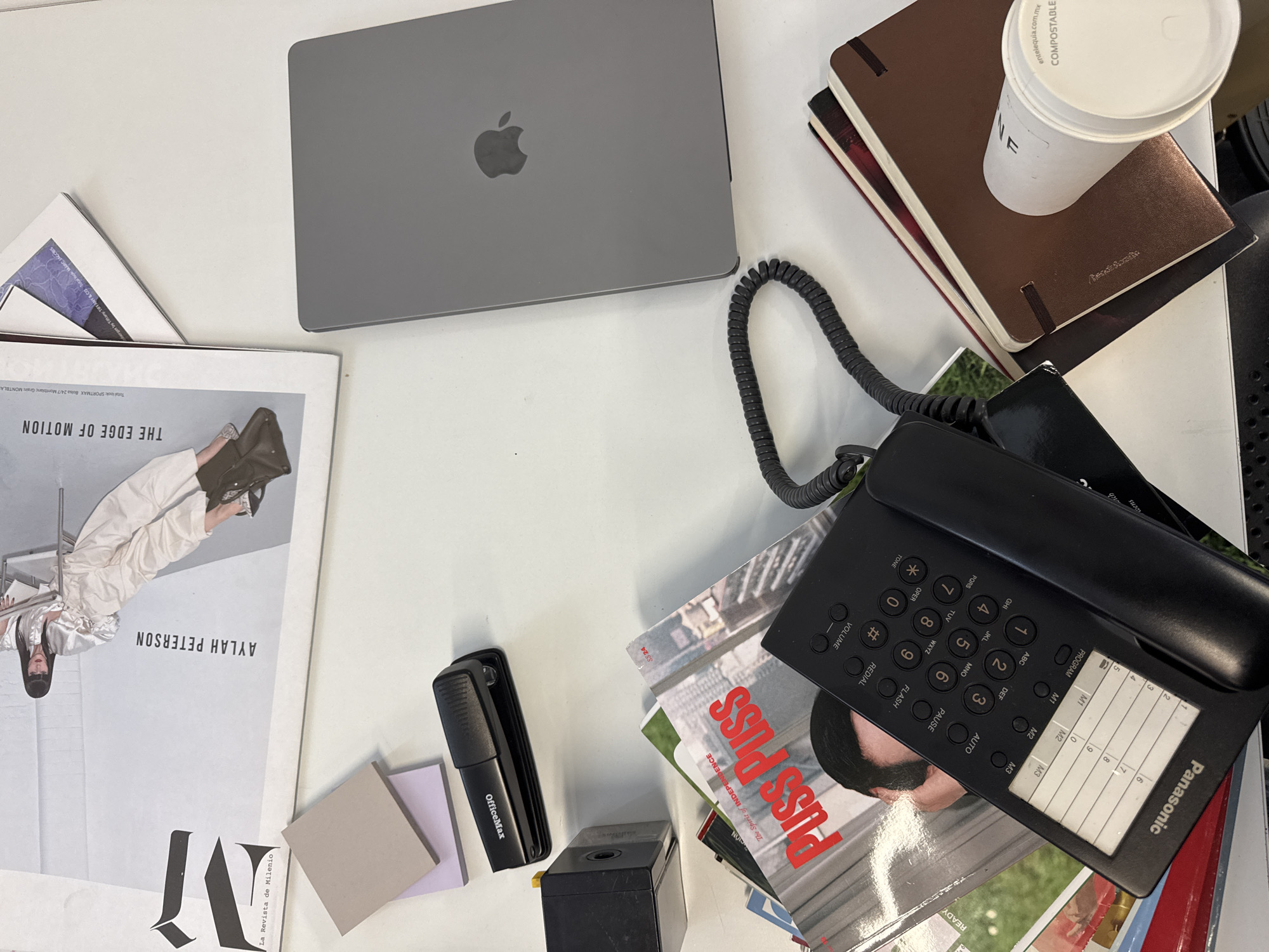 The image shows a cluttered desk. At the top left is a closed gray laptop with an Apple logo. Below the laptop is a stack of magazines, with the cover of "La Revista de Milenio" featuring "AYLAH PETERSON" and "THE EDGE OF MOTION" visible. Next to the magazine are sticky notes and an OfficeMax stapler.
To the top right is a stack of notebooks, and a paper cup labeled "COMPOSTABLE." A black telephone with a coiled cord and a Panasonic label is resting on another magazine stack near the bottom right. The magazine cover is titled "Puss Puss: The Spirit of INDEPENDENCE." A black box sits to the left of the telephone.
The location is Mexico City, Mexico, based on the "entelequia.com.mx" text on the paper cup and "La Revista de Milenio."