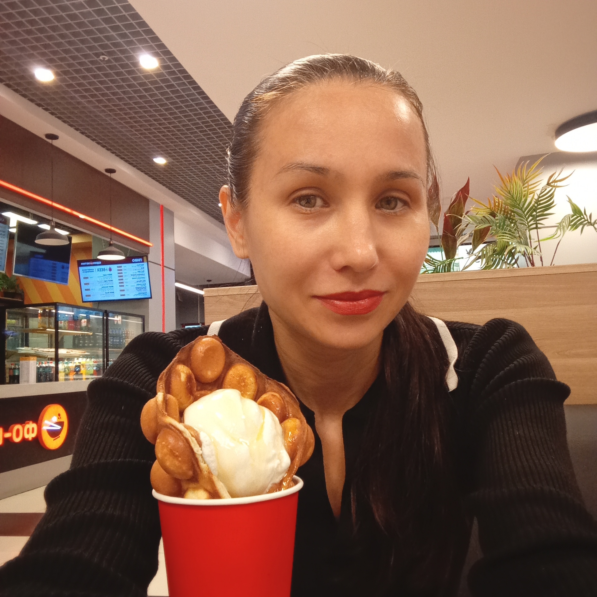 In Izhevsk, Russia, a woman with dark hair tied back and wearing a black top with white trim is holding a bubble waffle with ice cream in a red cup. She is looking at the camera, and her lips are painted red. In the background, there are signs with text in Cyrillic, fluorescent lighting, and a display case with drinks. The setting appears to be an indoor food establishment, possibly a cafe or kiosk.