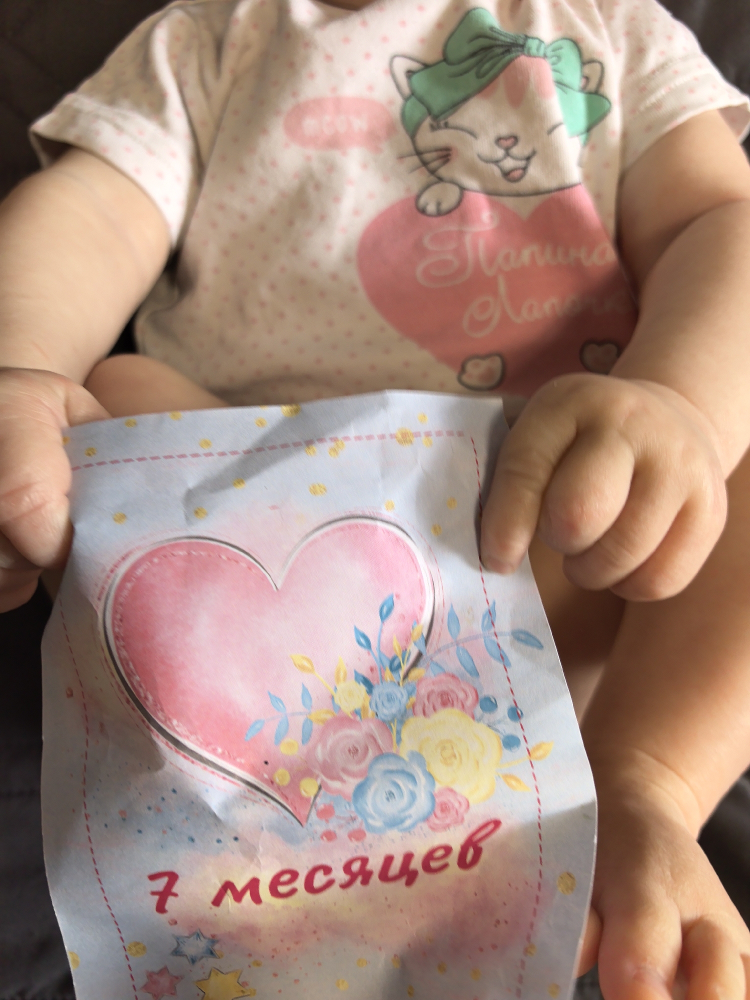 The media file is a close-up, vertical shot of a baby's torso and arms, holding a card. The baby is wearing a white t-shirt with small pink polka dots and a graphic of a cartoon cat with a green bow on its head, holding a large pink heart with text on it. The card in the baby's hands is light blue with yellow polka dots and decorative stitching. It features a large pink watercolor heart with a floral design. The visible text on the card reads "7 месяцев" (7 months) in bright pink cursive lettering. Below this, there are colorful star shapes. The lighting suggests it is daytime, and the setting appears to be indoors, likely a home environment in Фролы, Russia, given the context. The baby's hands are holding the card, and the focus is on the card and the baby's hands and chest. The overall mood is sweet and celebratory, likely marking a baby's 7-month milestone.