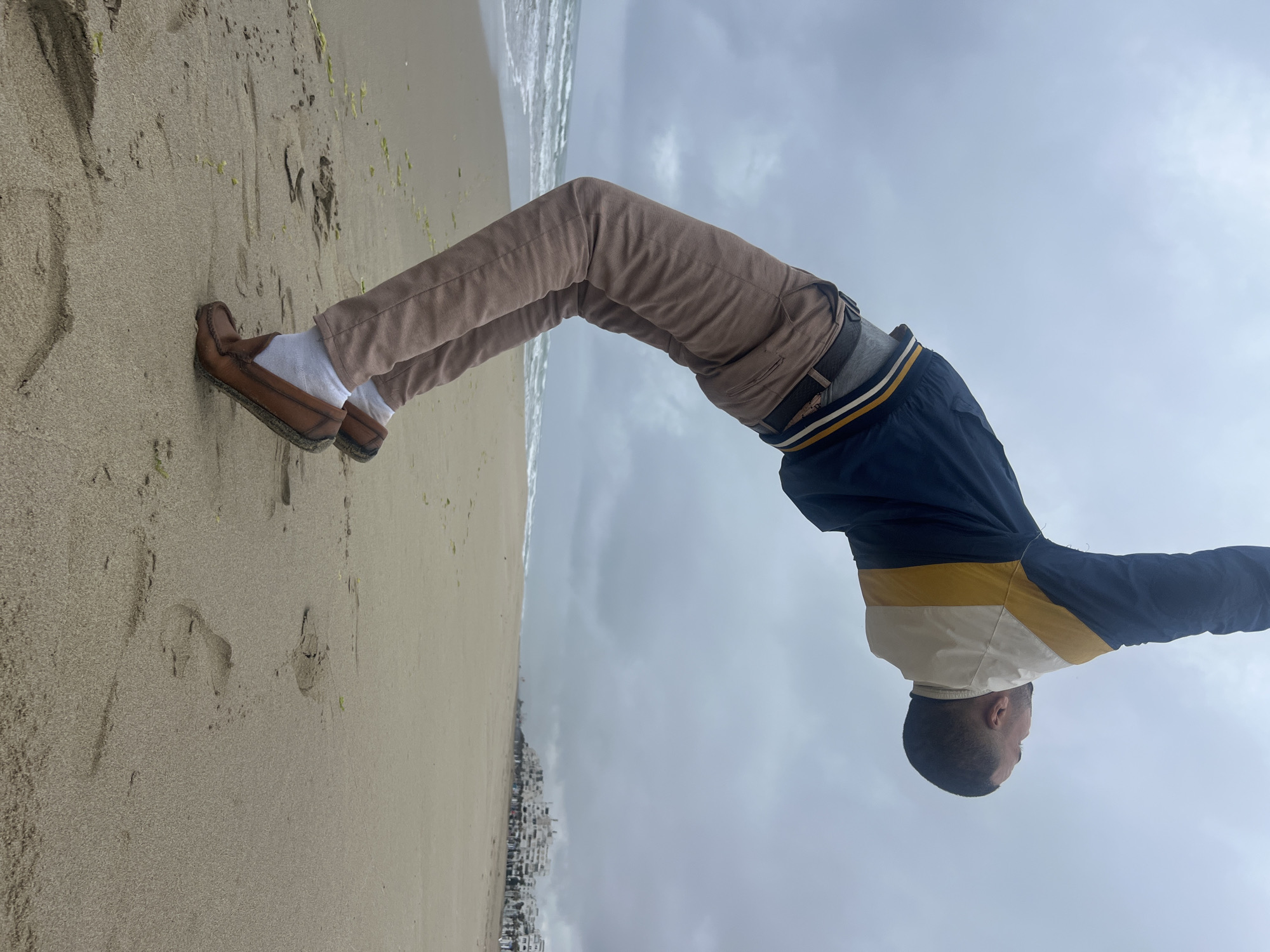 The image, rotated 90 degrees counter-clockwise from its natural orientation, depicts a person performing a backbend on a sandy beach in Martil, Morocco. The individual, appearing male with a shaved head, is wearing light brown trousers, white socks, and brown moccasin-style shoes. His upper body is covered by a distinctive jacket that is predominantly dark blue, with a section of cream and a mustard yellow stripe across the chest and shoulders.
The activity is a physical pose, specifically a backbend or bridge, performed on the soft, light-colored sand of the beach. The sand shows various textures, ripples, and some scattered green seaweed or debris. In the background, the ocean's gentle waves are visible, meeting the shore. Further along the horizon, a cluster of white buildings, characteristic of a coastal town or city, lines the distant landscape. The sky above is entirely overcast and cloudy, indicating a daytime scene without direct sunlight. No discernible text is visible on the person's attire or within the environment. The overall impression is one of physical expression set against a tranquil, cloudy beach environment.