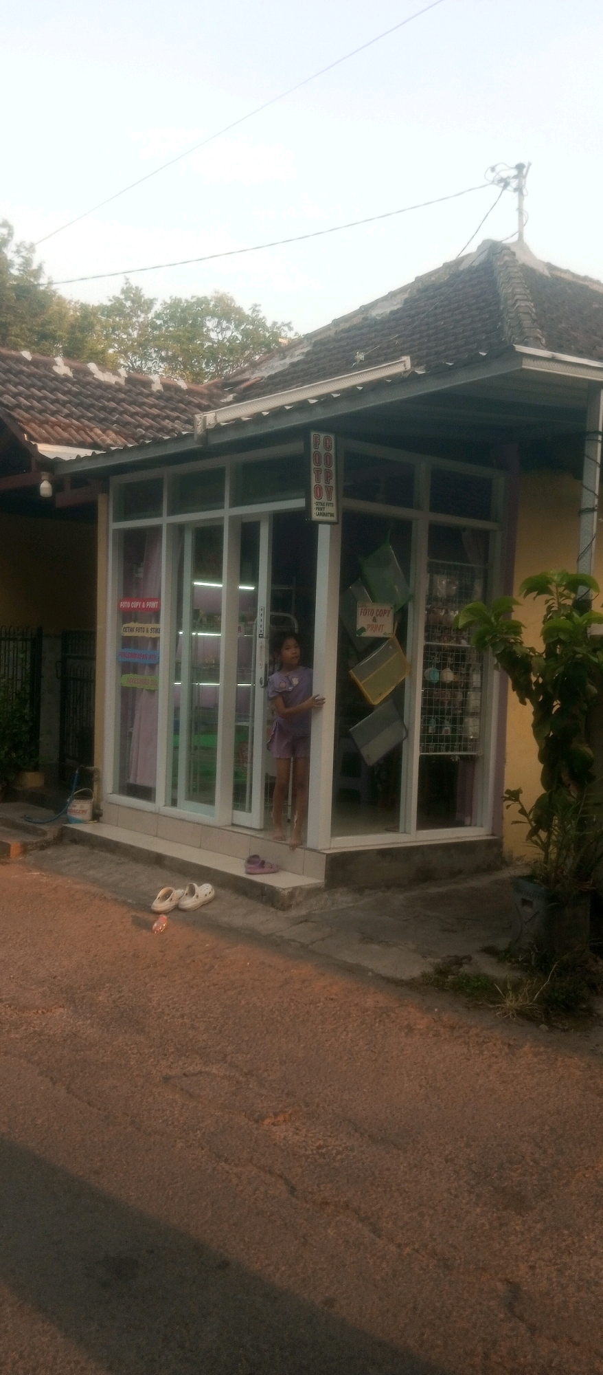 The image shows a small shop with a glass front, possibly a photo copy or printing service, located in Kediri City, Indonesia. A young girl in a purple dress stands partially in the doorway of the shop. Outside the shop, on the tiled step, are a pair of pink sandals and a pair of white sandals. The shop has signage that appears to indicate photo copy and printing services. Inside, shelves are visible displaying items, and there are some green and yellow folders hanging near the entrance. A tree with green leaves is visible to the right of the shop. The shop is situated on a paved road, with a shadow cast across it. The sky is bright, suggesting it is daytime.