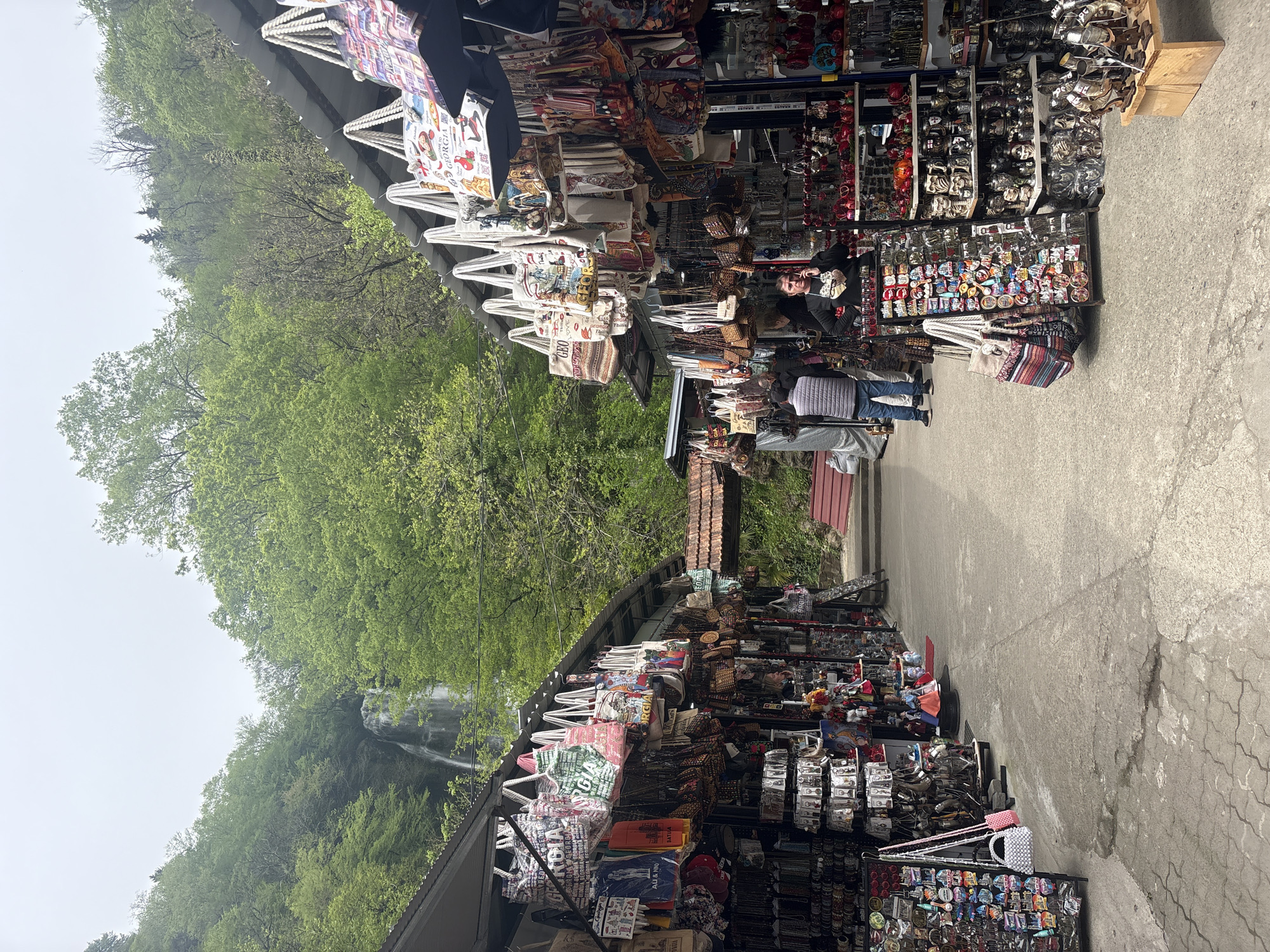 This image captures an outdoor souvenir market bustling with activity, set against a lush, tree-covered hillside in Kveda Makhuntseti, Georgia. Market stalls are arranged along both sides of a concrete path, displaying a wide array of merchandise. Tote bags, many prominently featuring the text "GEORGIA" in various fonts and designs, hang from the stall structures. Other items include numerous magnets, keychains, small ceramic statues, and other trinkets.
Several people are visible browsing the stalls. One person in a striped shirt appears to be looking at the items on a stand, while another individual wearing a light blue puffer vest stands further down the path. The scene conveys a casual, daytime shopping experience.
In the background, a dense, vibrant green forest covers a steep incline. A multi-tiered waterfall can be seen cascading down the hillside through the trees, partially obscured by the foliage, highlighting the natural beauty of the location. The sky is overcast or hazy, suggesting a cloudy day. Visible text on some merchandise includes "GEORGIA", "BATUMI", and possibly "ALL YOU NEED". The overall setting combines commercial activity with a striking natural landscape.