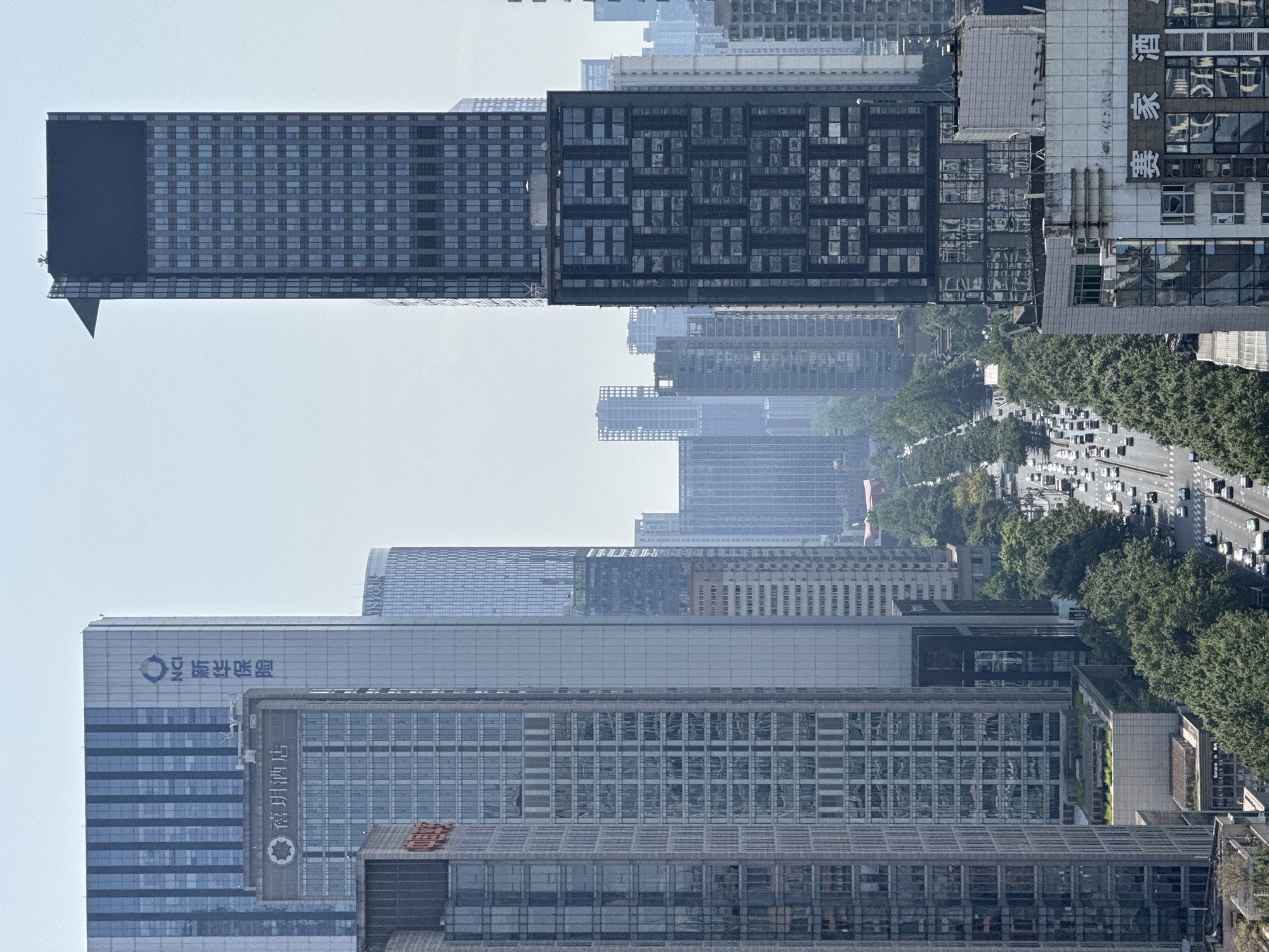 The image is a high-angle view of buildings in the Jinjiang District, China, with a focus on several modern high-rise buildings. The buildings are densely packed and vary in architectural style, with some featuring gridded facades and others with more sleek, modern designs. There is a street with cars and trees visible between the buildings. The sky is hazy. The image has been rotated counterclockwise.