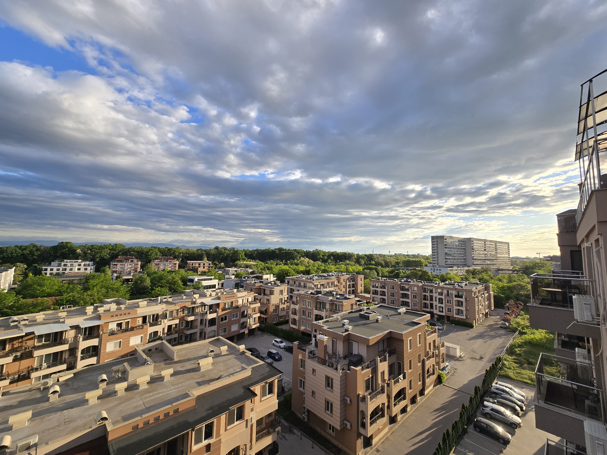 The image presents an elevated view over a residential area, likely on the outskirts of a city. Several mid-rise apartment buildings with a similar architectural style are visible. The buildings are constructed from light brown brick, featuring balconies and multiple windows. Parking lots are situated between the buildings, containing parked cars. In the background, a larger high-rise building is visible amidst green trees. The sky is filled with clouds, suggesting an overcast or partly cloudy day. The light indicates it may be late afternoon or early evening. The scene captures a quiet, suburban neighborhood with its characteristic architecture, parking arrangements, and surrounding greenery.