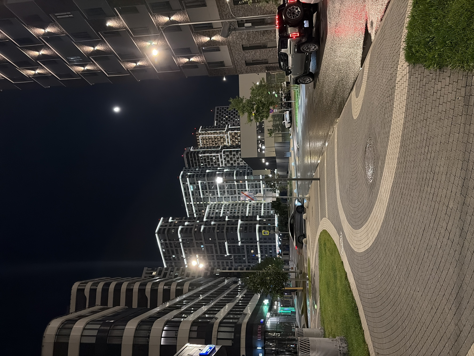 The image depicts a nighttime scene in Moscow, Russia, specifically in the residential complex "Spires" (Шпилей) or a similar modern development. The perspective is from a paved sidewalk with a curved, two-toned brick pattern, bordered by a grassy area on the left and a street on the right.
Dominating the background are several multi-story residential buildings, illuminated from within and with external accent lighting that outlines their modern architectural features. The buildings are characterized by their geometric shapes and large windows, indicative of contemporary urban design. A bright moon is visible in the dark night sky, positioned above the buildings.
In the foreground and midground, the street is wet, suggesting recent rain, and reflects the lights from the buildings and streetlights. Two cars are visible on the street. On the right, a silver Hummer EV SUV with its taillights illuminated is parked or stopped, facing towards the left. Further down the street, a darker colored sedan is in motion, heading away from the viewer and towards the illuminated buildings in the distance. Streetlights are present, casting a warm glow on the surroundings.
The overall impression is one of a well-maintained, modern urban environment at night, with the wet streets adding a reflective and somewhat dramatic quality to the scene. There are no people visible in the frame.