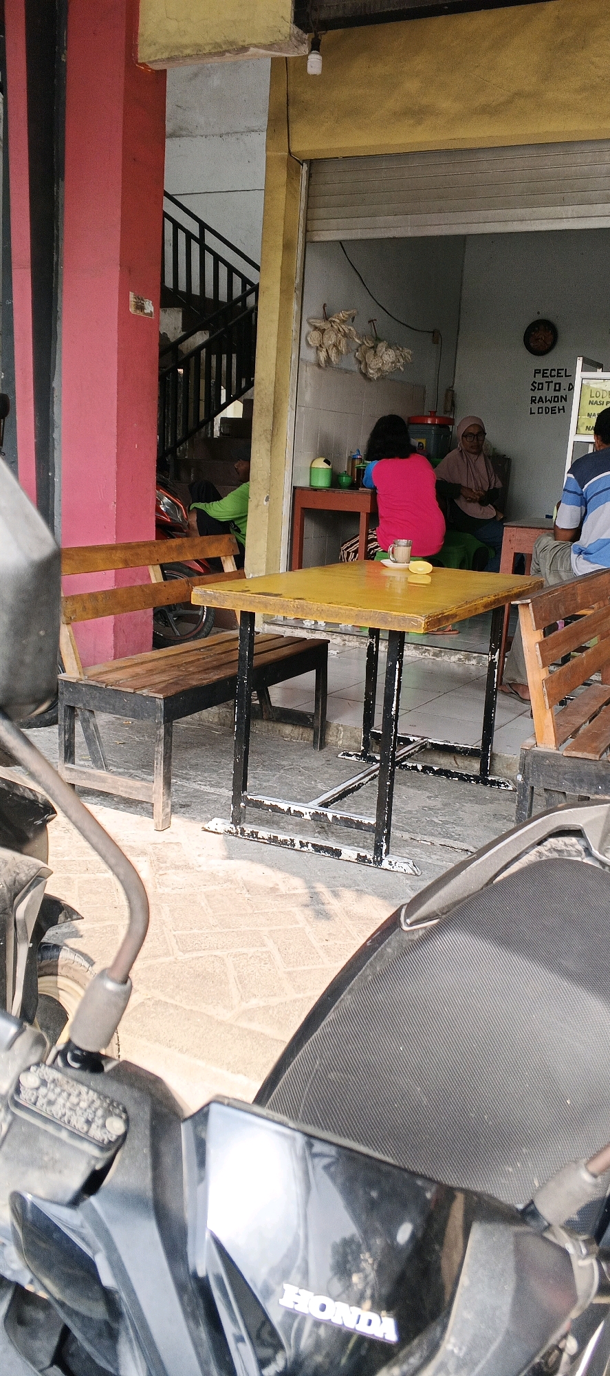 The image captures an outdoor seating area of a food stall in Karanggeneng, Indonesia. A yellow table with black metal legs is positioned centrally, with a wooden bench to its left and a wooden chair to its right. On the table, a cup of coffee and two wedges of fruit are visible. In the background, through an open doorway, three individuals are seated. One woman in a pink shirt sits closest to the camera, facing away. To her left, another woman wearing a headscarf and glasses is seated. A third person, partially visible, is seated to the right. The stall's exterior features red and yellow paint. A staircase is visible to the left, and a red motorcycle is parked near it. The ground is paved with light-colored rectangular tiles. The foreground of the image is dominated by a portion of a black motorcycle.