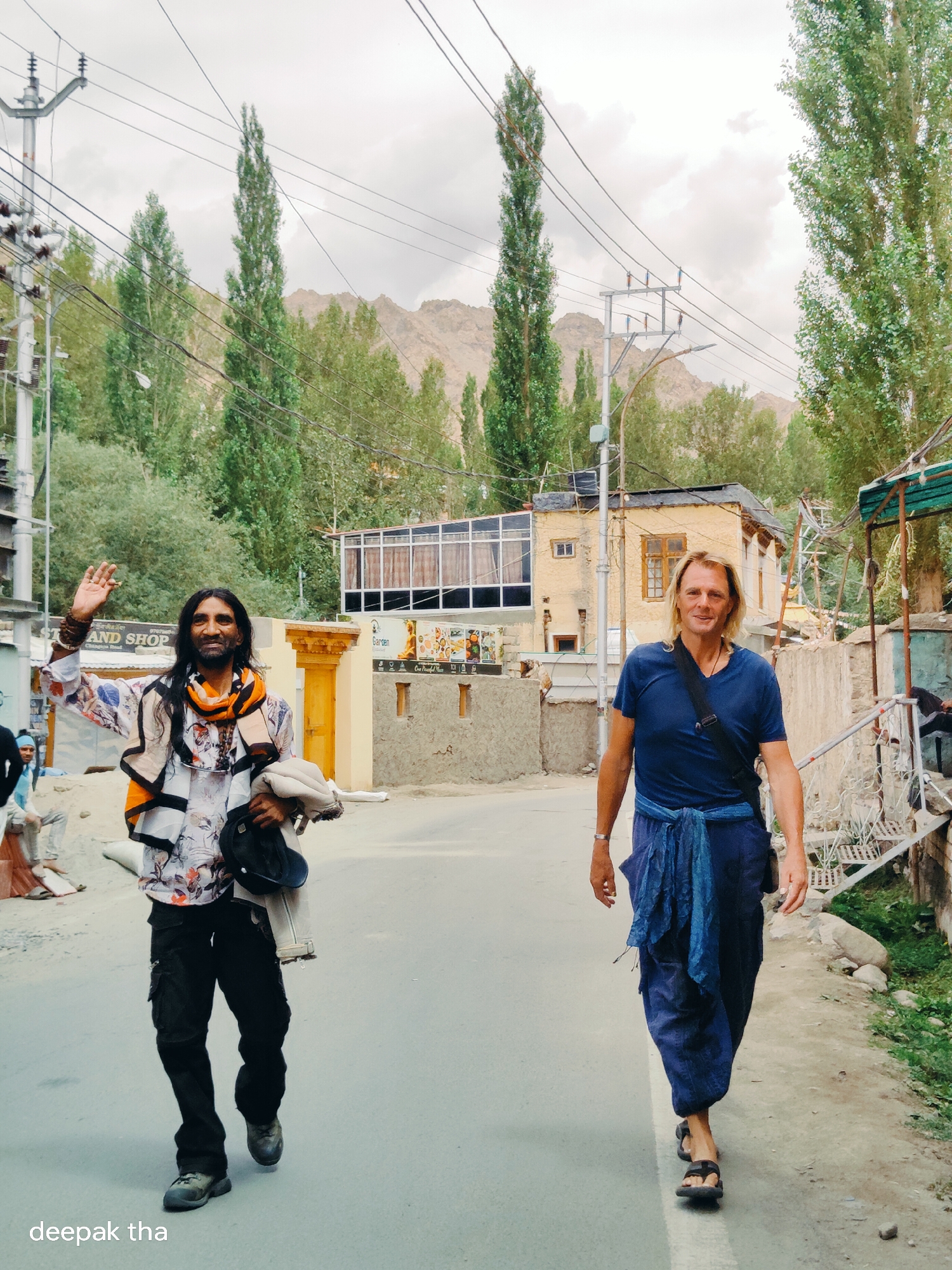 The image shows two men walking on a street in the city of Leh, India. The man on the left is waving and appears to be a local resident, dressed in traditional attire and holding a jacket and hat. He has long dark hair and is wearing a vibrantly colored scarf. The man on the right is a foreigner with blond hair, wearing a blue t-shirt, loose blue pants tied at the waist with a blue sash, and sandals. He has a dark strap across his chest. In the background, there are buildings with traditional Ladakhi architecture, trees, and mountains. Utility poles and wires are visible, indicating an urban setting. The street is paved, and there is some vegetation and rocks along the side.