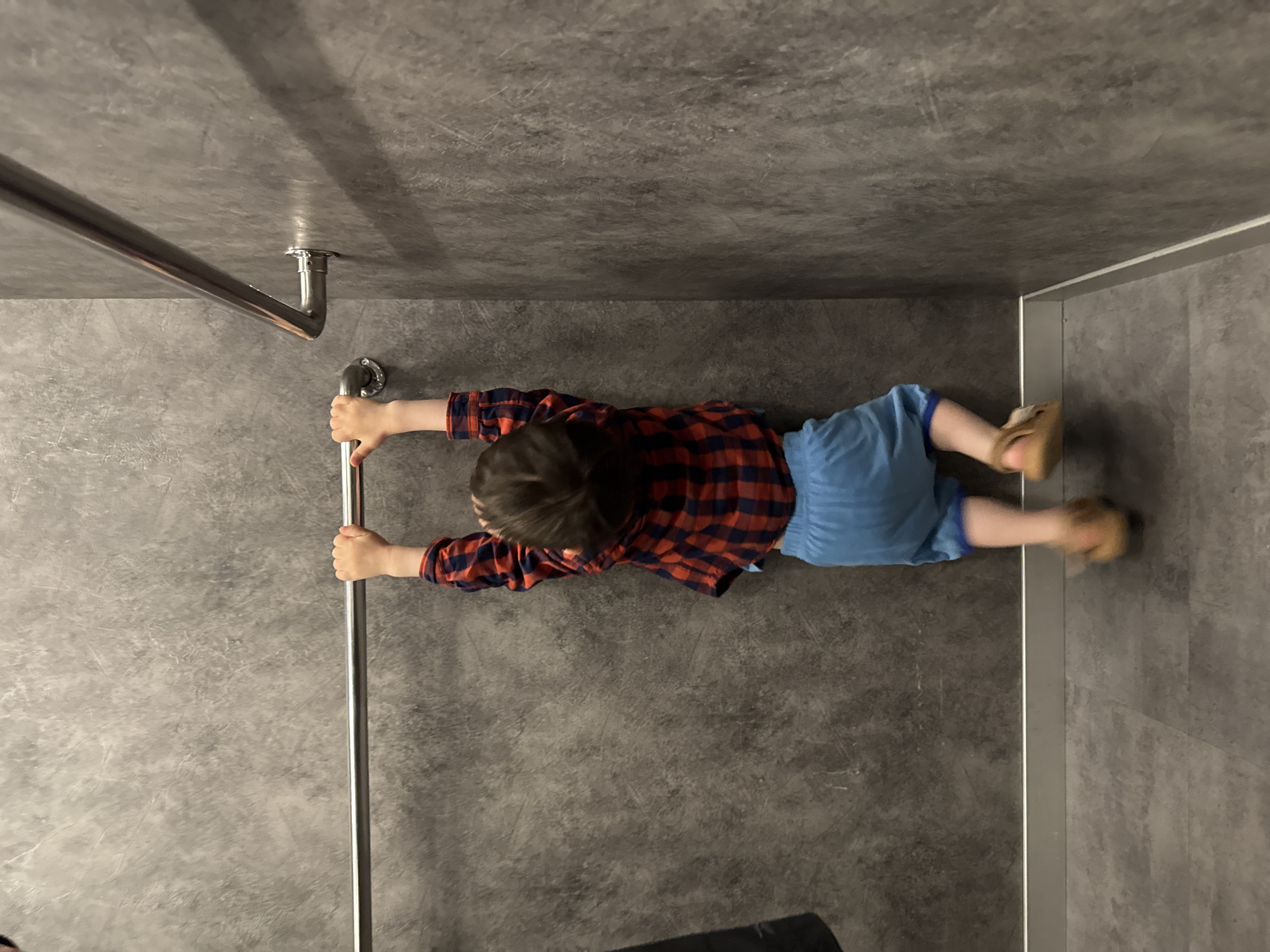 A child is lying on their stomach in what appears to be a bathroom stall. They are holding onto a silver metal grab bar on the left side of the frame. The child is wearing a red and blue plaid shirt, blue shorts, and tan shoes. The walls and floor are gray with a textured finish. A silver trim runs along the right wall. The location is unknown and not clearly evident in the image. The presence of Sochi, Russia, is undetermined.