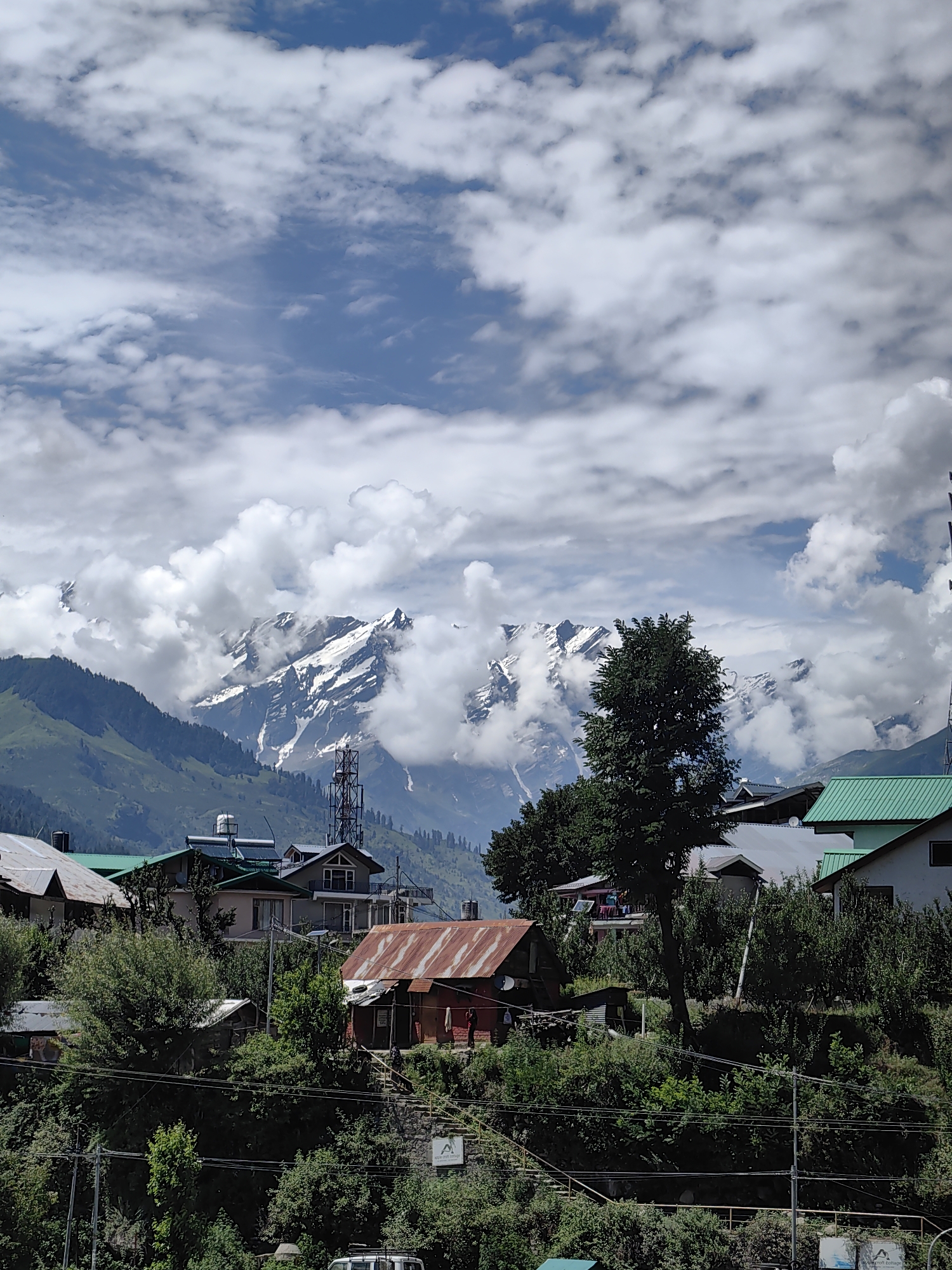 The image depicts a village nestled in the foothills of snow-capped mountains, likely in Siumsa, India, given the context. The foreground features a collection of houses with various roofing materials, including rusted corrugated iron and green metal sheeting. Lush green trees and dense foliage surround the buildings, with several power lines crisscrossing the scene.
In the midground, a prominent telecommunications tower stands tall, juxtaposed against the natural landscape. Beyond the village, rolling green hills rise, transitioning into the majestic mountain range. These mountains are partially obscured by dramatic, white clouds, but their snow-covered peaks are visible, creating a striking contrast with the blue sky. The overall atmosphere is one of a remote, mountainous settlement under a partly cloudy sky.