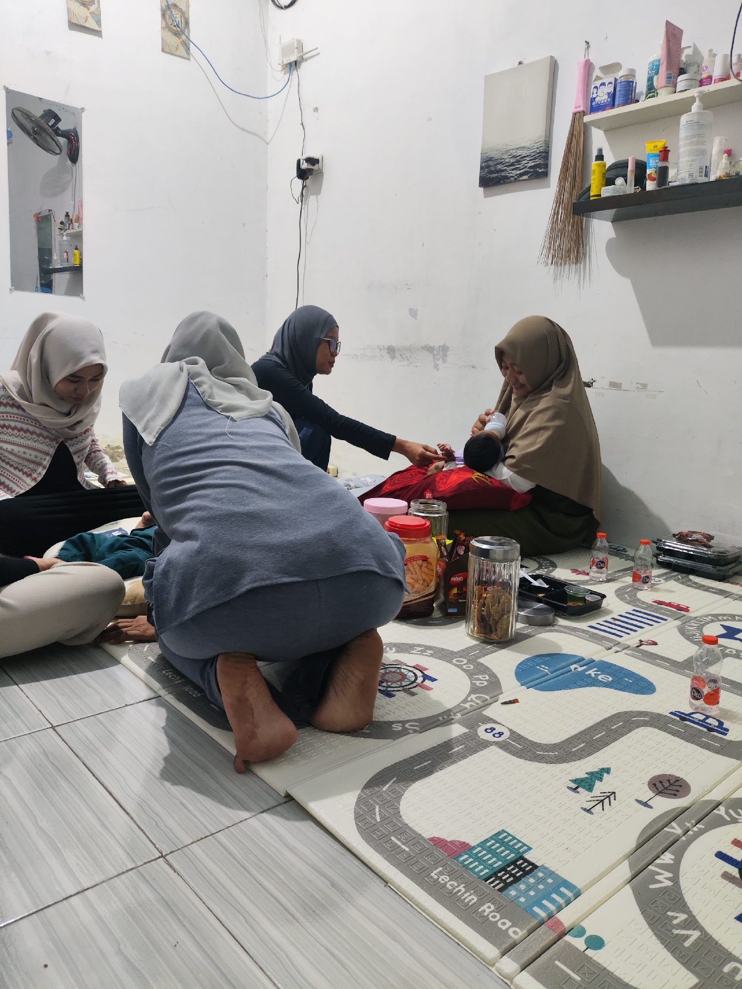 In a room with white walls and tiled flooring, four people are gathered on the floor. A baby is being held and fed a bottle by a woman wearing a light brown hijab. To her left, another woman in a grey hijab looks on. A third woman, also in a light-colored hijab, is seated behind them. The fourth individual, facing away from the camera, is positioned in the foreground, wearing a grey outfit. The floor is covered with a play mat depicting roads and buildings. Scattered around are food containers, bottles, and other miscellaneous items. On the wall, a fan is mounted, and shelves hold various products. The scene appears to be in the city of Prague, Indonesia.