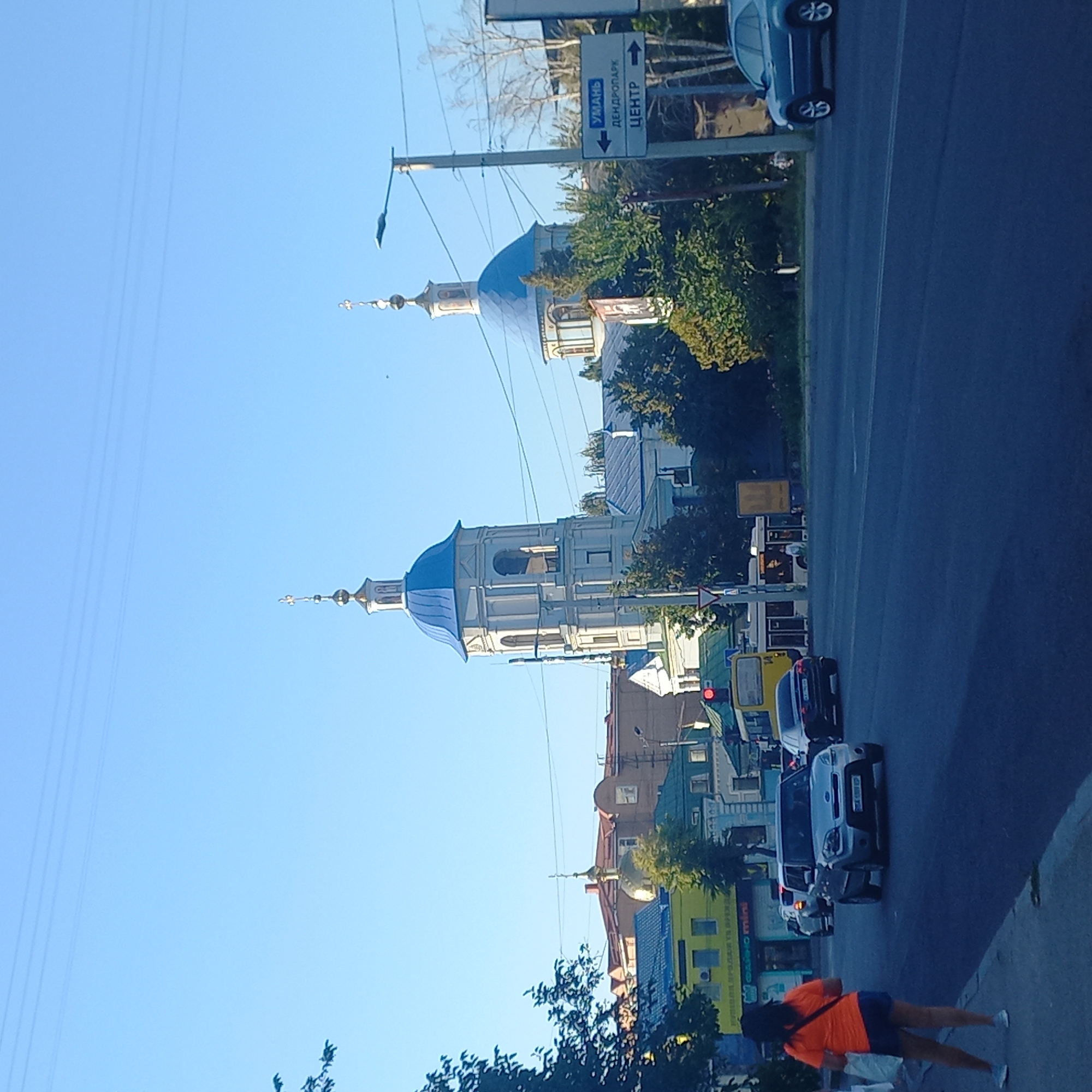 The image depicts a street scene in Kropyvnytskyi, Ukraine, likely during daylight hours due to the bright sky and shadows. A woman in an orange t-shirt and blue shorts stands on a sidewalk facing away from the camera. She is carrying a white bag. The street is paved and appears to be a multi-lane road. Several cars are present on the road, including a silver Kia Soul in the foreground. Traffic lights are visible, one red and one green. In the background, there are buildings, including a church with a prominent blue dome and a bell tower, and another building with a golden dome. Trees and vegetation are scattered throughout the scene. A street sign is visible on a pole with directional arrows and text in Cyrillic, indicating "УМАНЬ", "ДЕНДРОПАРК", and "ЦЕНТР". Power lines are suspended overhead.