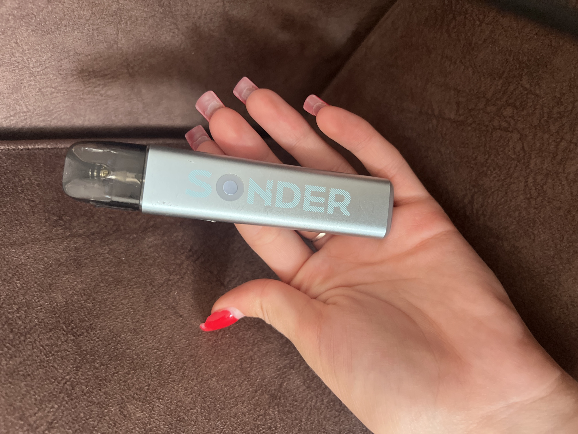 A person's hand, with bright red nail polish on the thumb and pale pink acrylic nails on the other fingers, holds a light blue colored vape pen with the word "SONDER" printed in light blue lettering on its side. The vape pen has a dark translucent cartridge at the top. The background is a textured brown surface, possibly upholstery. The image does not contain any identifiable landmarks or clues that would indicate its location in the city of Sheptytskyi, Ukraine.