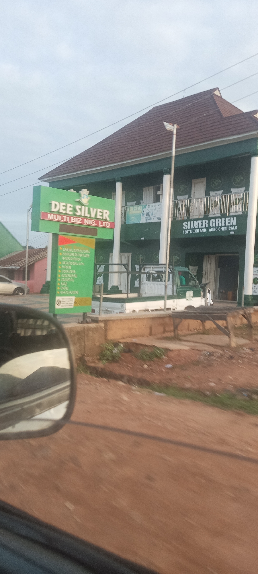The image, likely taken from a moving vehicle, shows a business complex in Mangu Town, Nigeria. The building is painted green and has two visible signs: one for "DEE SILVER MULTI BIZ NIG. LTD" and another for "SILVER GREEN FERTILIZER AND AGRO CHEMICALS." A white and green truck is parked in front of the building. The surrounding environment appears to be a roadside with some vegetation and dirt. In the lower left corner, a car mirror is visible.