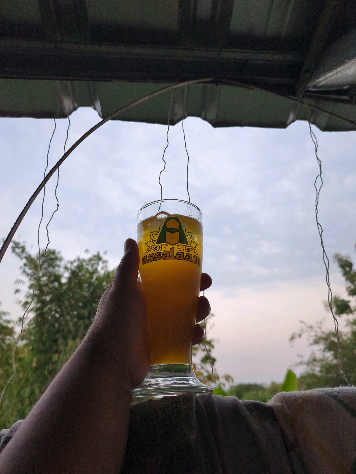 A hand holds a glass of golden liquid, possibly beer or juice, against an overcast sky. The glass has a logo featuring a green crescent and star above decorative text that reads "assalaam". The scene appears to be outdoors, with lush green trees and foliage visible in the background. The foreground is occupied by a dark, textured surface that could be fabric. The overall impression is one of a relaxed moment in a natural setting, possibly in or near Sindon, Indonesia.