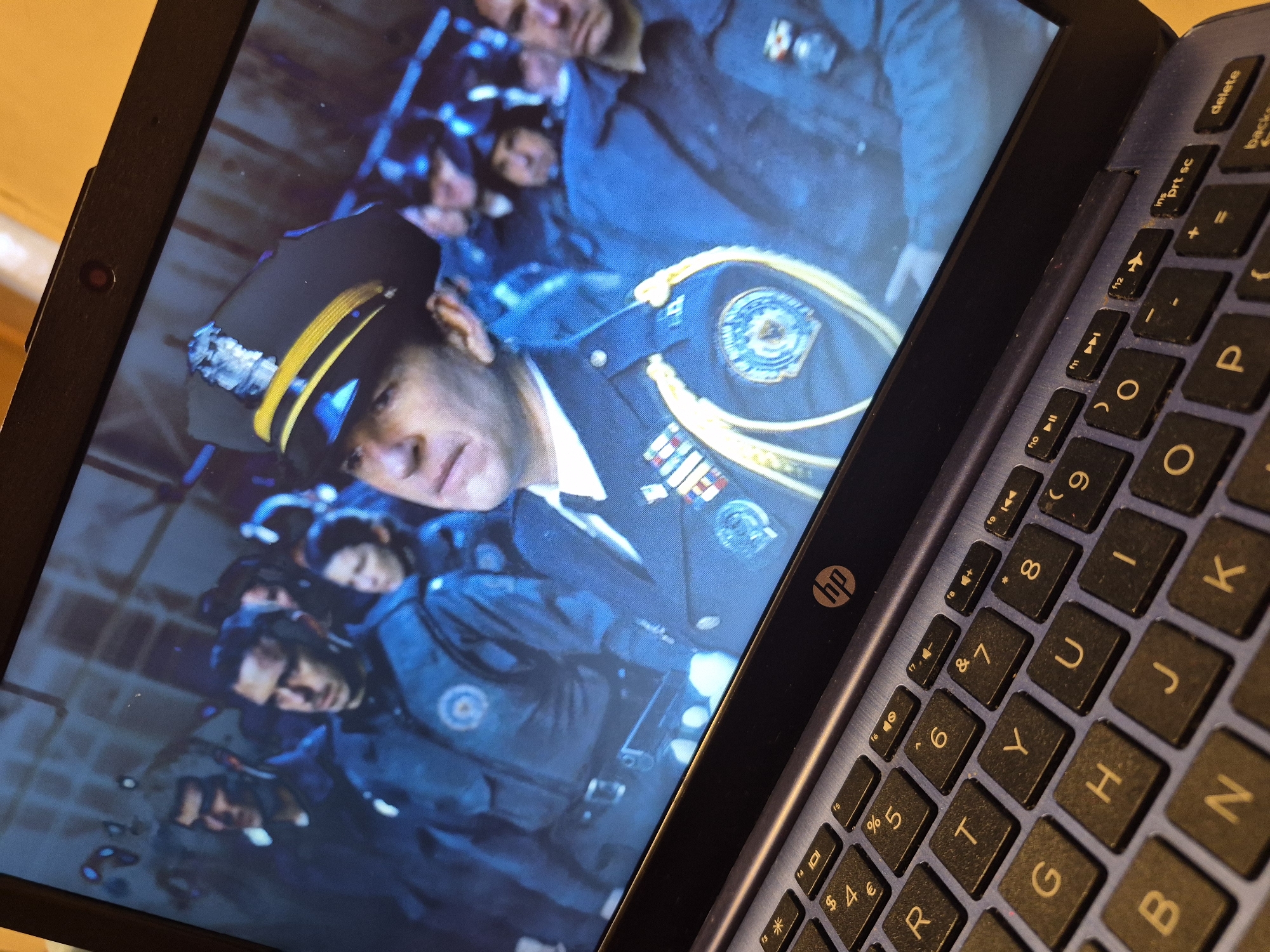 The image is a close-up, angled shot of a laptop screen displaying a still frame from a video, placed on a keyboard. The video content shows a group of people in uniform, appearing to be police officers, gathered in what looks like an indoor, dimly lit setting with a brick or tiled background. The central figure in the foreground is a man wearing a police officer's uniform, including a cap with gold trim and a badge, and several medals on his chest. He is looking slightly to his left. Behind him, other officers in similar uniforms are visible. The laptop's keyboard is in the foreground, showing various keys, including letters, numbers, and symbols, and the HP logo is visible on the laptop's bezel. The overall lighting suggests a scene from a movie or television show. There is no information to suggest this image or its content is located in Sandwell, United Kingdom.