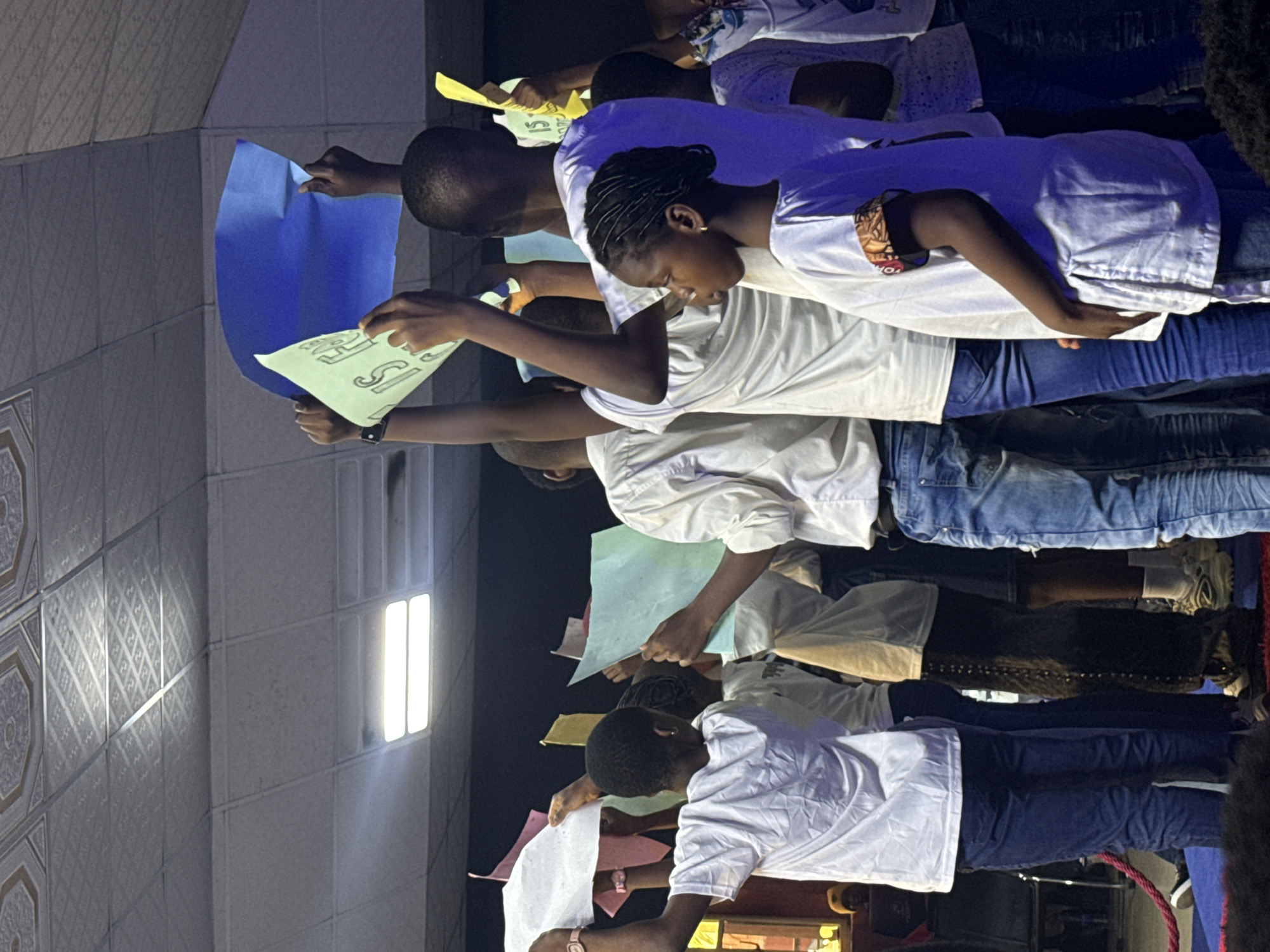 A group of approximately seven people, mostly teenagers and young adults, stand in a row holding up pieces of colored paper and white signs with lettering on them. The people in the group are wearing white t-shirts and jeans. The image was taken indoors; the background indicates a room illuminated by two white fluorescent light panels. The location is Nkpor, Nigeria.