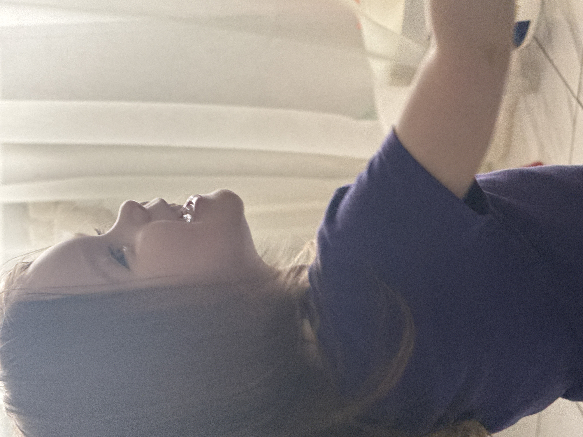 A woman with light skin and long brown hair lies on her back. She is wearing a dark blue t-shirt, and her head is tilted back, exposing her neck and chin. Her mouth is slightly open in a smile or laugh. The background consists of blurred, neutral-toned surfaces, possibly walls or fabric, with indistinct lighting. The location is Unknown, Unknown.