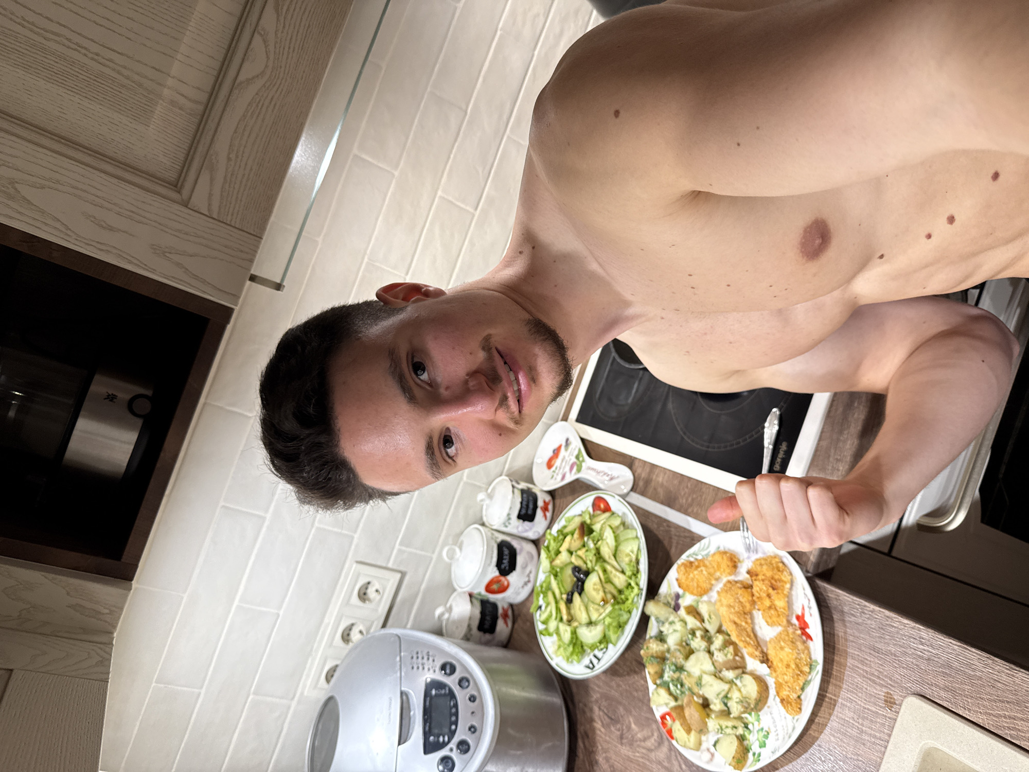 A man with short dark hair and a goatee is standing in a kitchen, shirtless, holding a fork in his right hand, angled towards the camera. He appears to be taking a selfie. On the kitchen counter in front of him are two plates: one with a green salad of cucumbers and olives and another with fried chicken tenders and potato salad. Behind the plates, there are three white ceramic jars labeled with the word "SALT" stacked on top of each other and a ceramic spoon rest. To the left, there is a bread-making machine, electrical outlets, and white tiled walls with wooden kitchen cabinets. Behind him, there is a black stovetop and a stainless steel oven. The location appears to be a kitchen in a private residence. There is no specific visual indication of Sofia, Bulgaria.