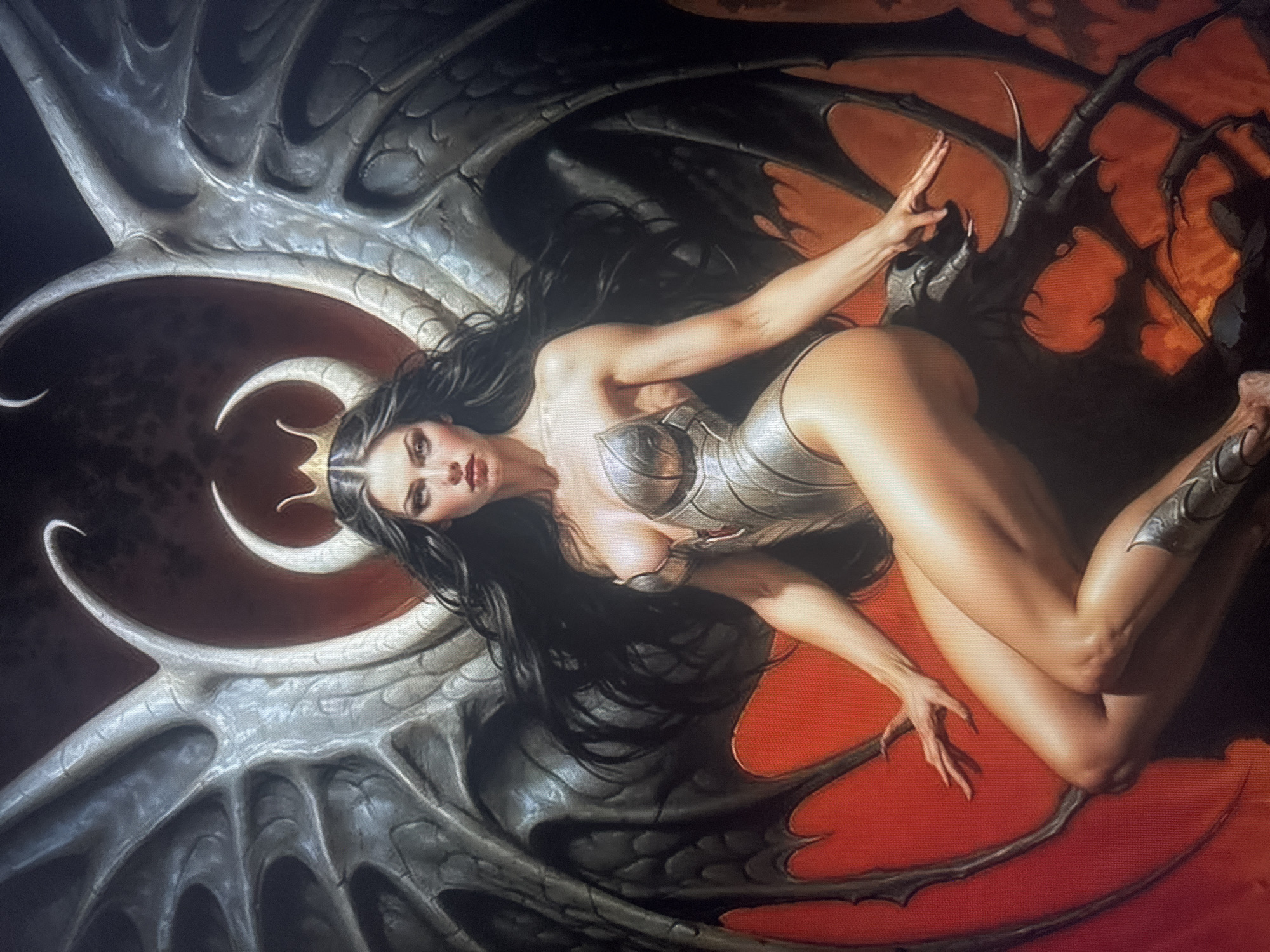 The image depicts a fantasy illustration of a female figure with large, dark, skeletal wings and curved horns. She is wearing a metallic, form-fitting outfit and a crown. The background is dark with some red and orange hues. The figure is seated in a dramatic pose. The image does not contain any recognizable elements or landmarks that would allow for the determination of a location in the city of Dondușeni, Moldova.