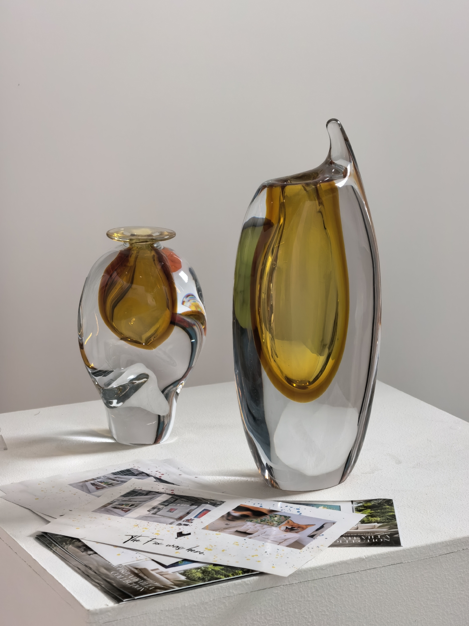 A still life photograph features two abstract glass vases displayed on a white surface. The vase on the left is shorter and wider, with a clear outer layer and an inner chamber filled with amber and swirled colors. The vase on the right is taller and more elongated, with a pointed top and a similar amber-colored interior within a clear glass exterior. In front of the vases, several printed sheets of paper are arranged, displaying images of interiors and landscapes, some with scattered yellow and blue flecks. The background is a plain, light-colored wall. The lighting is soft and diffused, casting subtle shadows. There is no indication of the city of Ko Samui District, Thailand in this image.