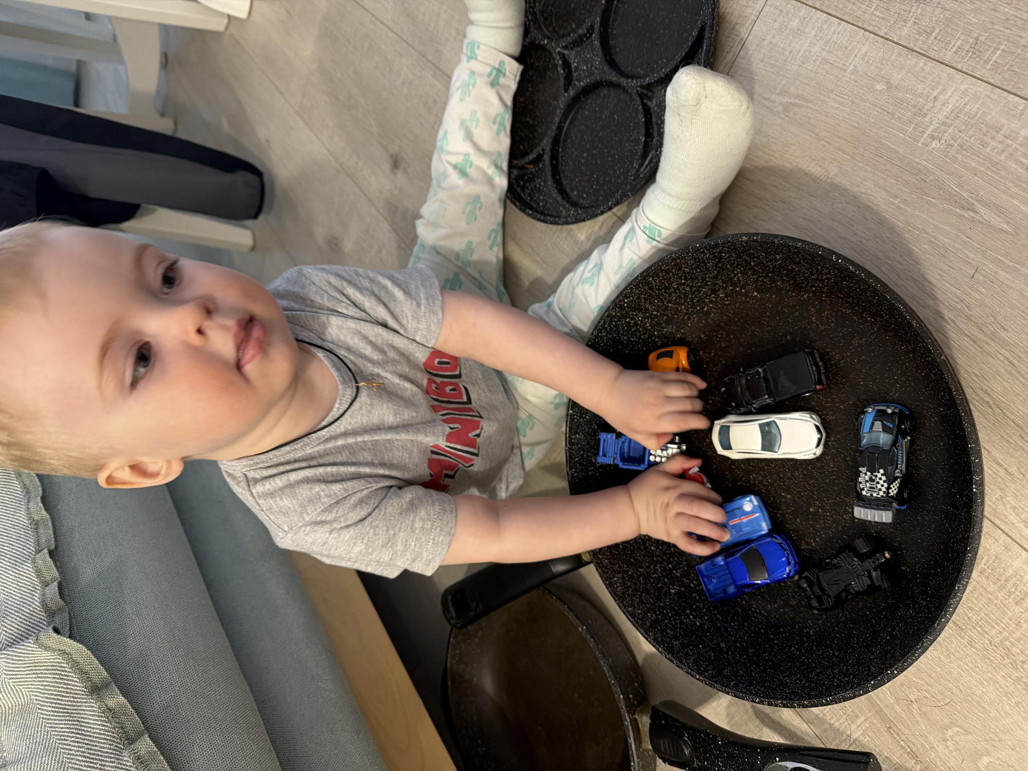 A child, approximately one year old, sits on a light-colored wood-grained floor. The child is wearing a gray t-shirt with a red and white graphic, light-colored pants patterned with green cacti, and white socks. The child is surrounded by kitchenware, including a large black frying pan filled with toy cars of various colors and models, and a smaller black pancake pan positioned behind the child's legs. The child is holding the toy cars in the frying pan. Part of a light blue upholstered couch is visible in the lower left corner. Based on the general domestic setting and lack of recognizable landmarks, it is not possible to confirm the location in the city of Votkinsk, Russia.