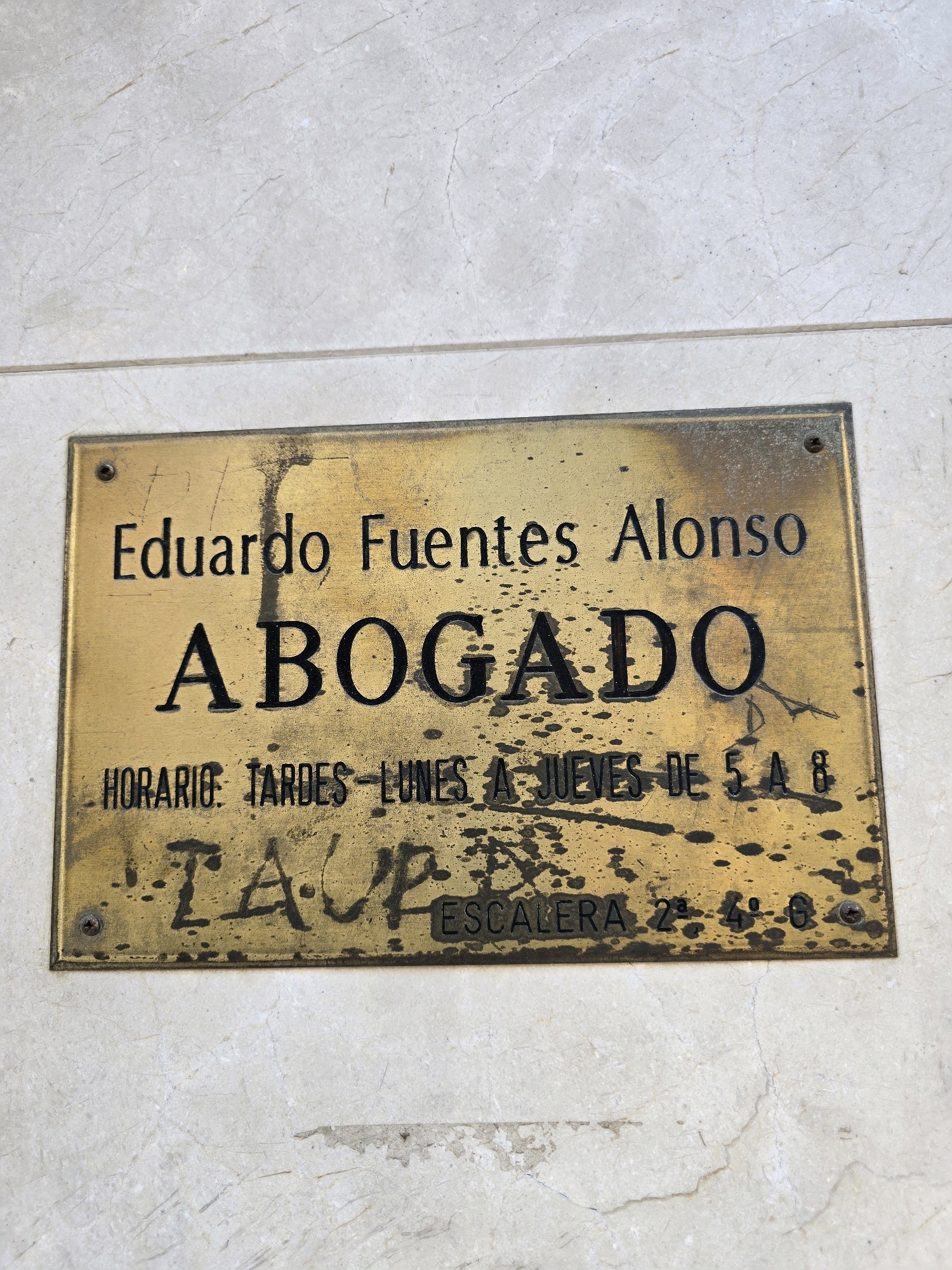 The image shows a brass plaque mounted on a marble wall. The plaque is inscribed with text in black lettering. The top line reads "Eduardo Fuentes Alonso". Below this, in larger letters, is the word "ABOGADO", which translates to "lawyer" in Spanish. The third line of text details the office hours: "HORARIO: TARDES - LUNES A JUEVES DE 5 A 8". This means "Hours: Afternoons - Monday to Thursday from 5 to 8". Below this, a scribbled graffiti in black spray paint covers part of the plaque. The graffiti consists of an illegible word followed by "ESCALERA 2 ª - 4º - 6º", which indicates "Staircase 2nd - 4th - 6th". The plaque is slightly tarnished and appears to be old. The wall it is mounted on is light-colored marble with subtle veining. There are no people visible in the image. The plaque is located in Jaén, Spain, as indicated by the text on the plaque itself.