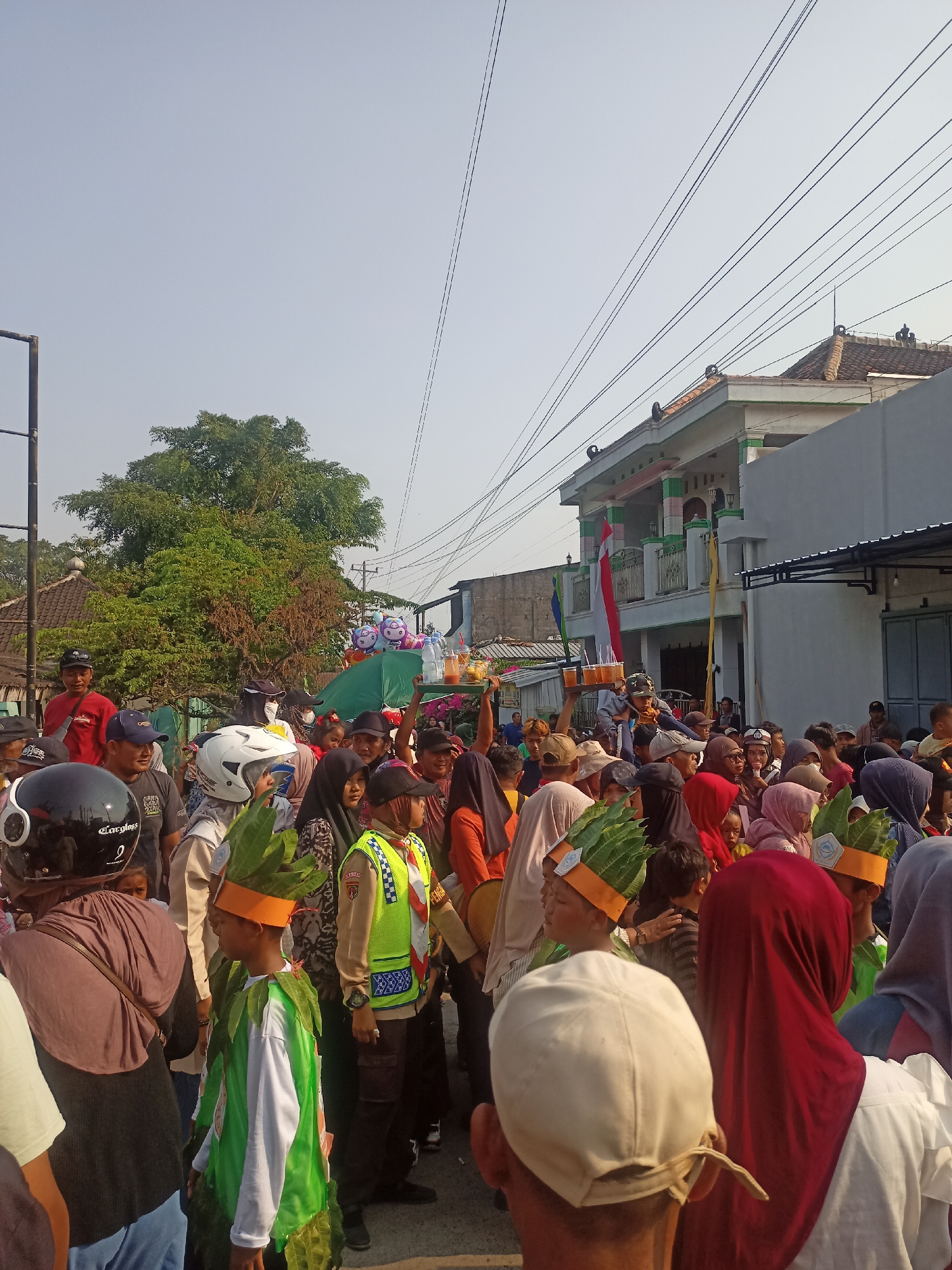 A dense crowd of people, predominantly adults with a few children, are gathered outdoors on a street in Gemolong, Indonesia. The scene appears to be a procession or parade, with some individuals wearing elaborate costumes. One child in the foreground is dressed in a green leafy outfit with a feathered headdress, while another child also wears a similar costume. A person in a bright yellow and blue safety vest, likely a traffic controller or event staff, stands prominently. Other people are dressed in everyday clothing, with many wearing head coverings, suggesting a mix of participants and onlookers. Some individuals are holding trays of drinks, and a few balloons are visible in the background. The environment includes a building with a traditional tiled roof and white exterior, suggesting a residential or commercial area. Overhead power lines crisscross the sky, indicating an urban or semi-urban setting. The sky is clear and the lighting suggests it is daytime, likely morning or afternoon.