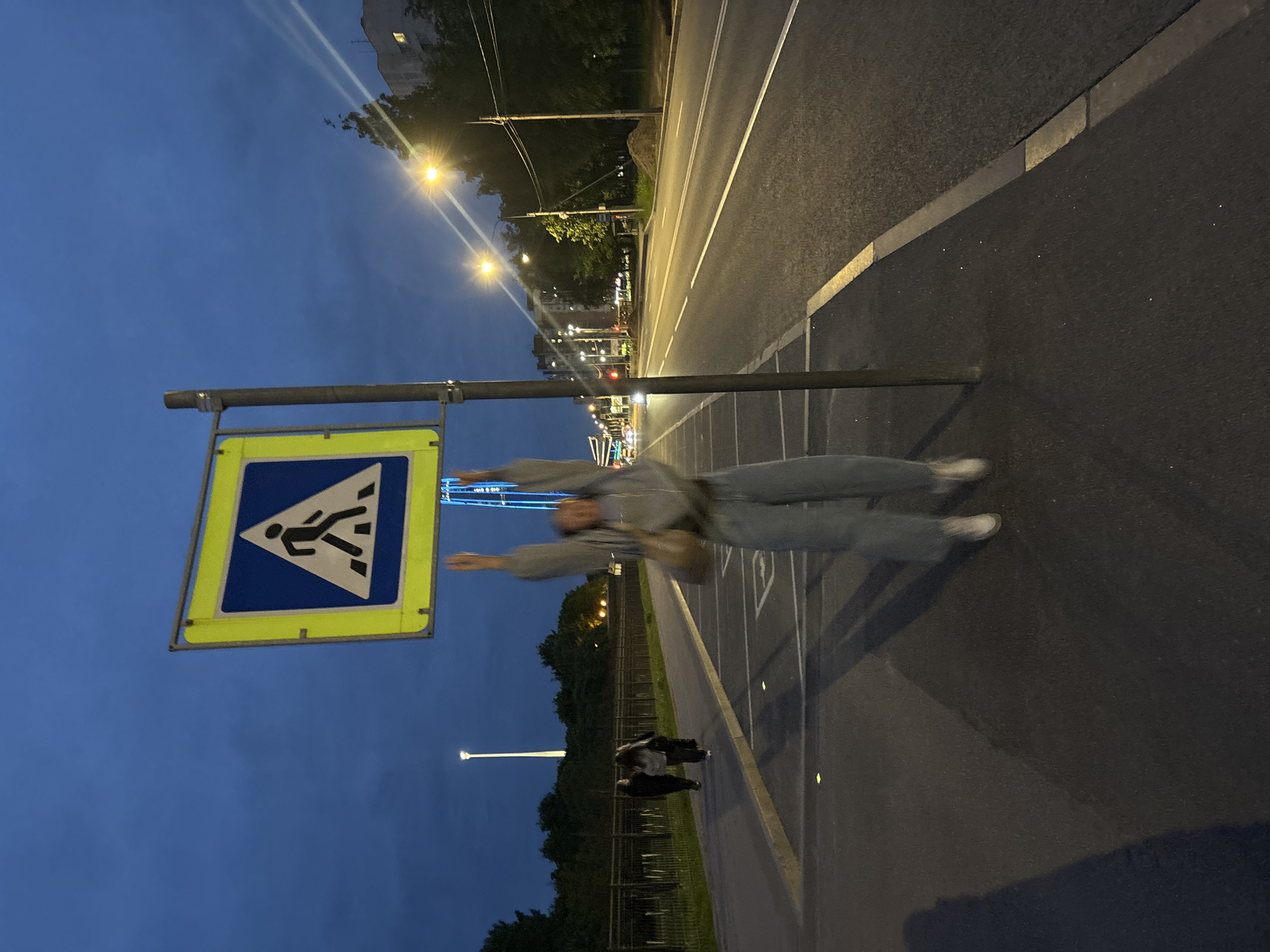 A medium shot captures a person in motion on a street in what appears to be Saint Petersburg, Russia. A pedestrian crossing sign with a yellow border and a blue symbol of a walking person is positioned on the left side of the frame. To the right of the sign, a blurred figure of a person wearing light-colored clothing is caught mid-stride. The sidewalk and street stretch out into the distance, illuminated by streetlights. In the background, other individuals are visible, and a tall, slender tower can be seen. The sky above is a gradient of blue, suggesting either early morning or late evening.