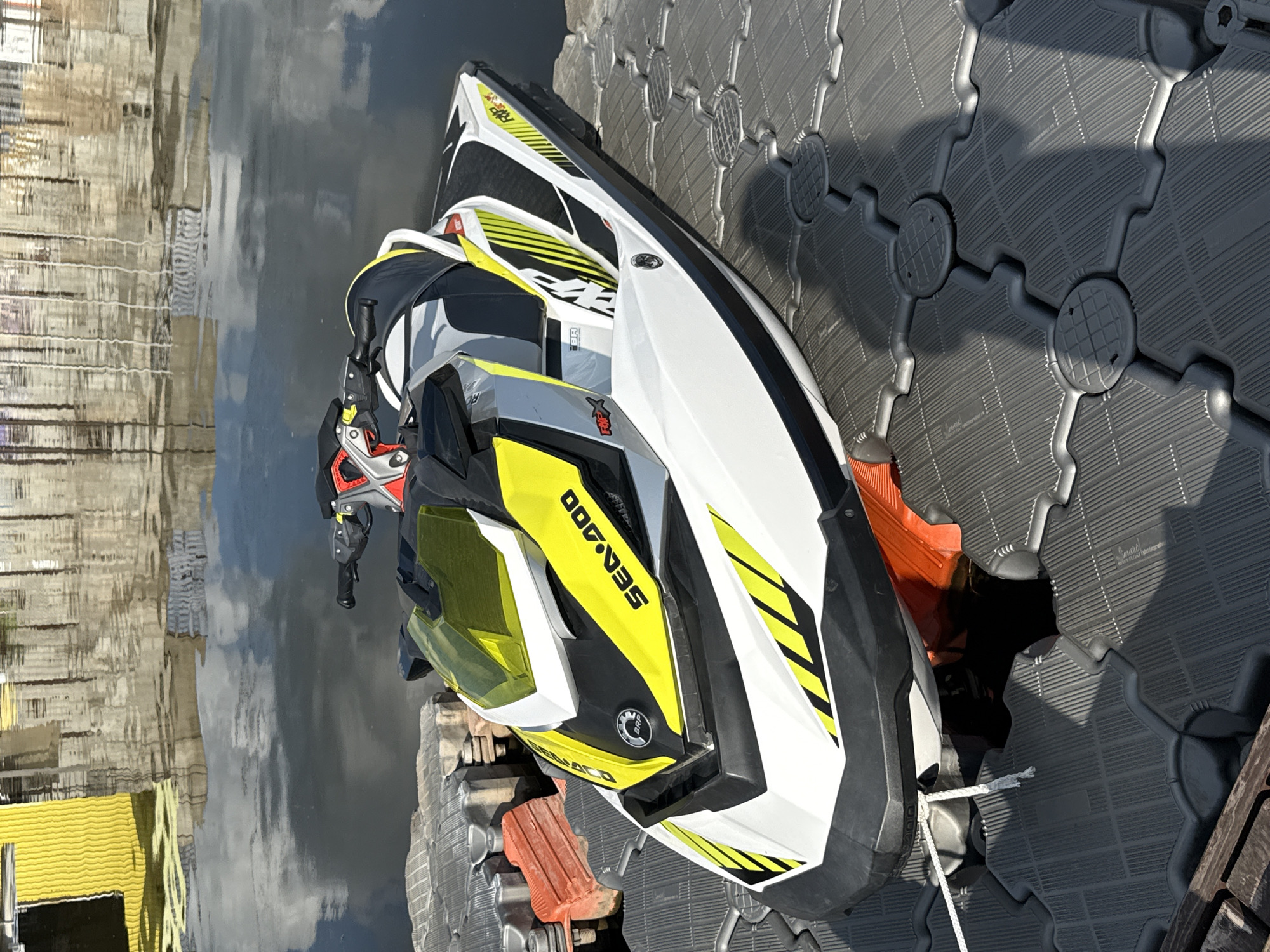 A white and yellow Sea-Doo RXP-X jet ski is parked on a gray modular floating dock. The jet ski is positioned at an angle, facing slightly to the right. It has black accents and yellow graphic stripes. The handlebars and front section with a yellow windshield are visible. A white rope is attached to the jet ski and runs down towards the dock.
The dock is made of interlocking gray plastic modules, some of which have circular raised elements. Red boat fenders are visible on the left side of the dock, near the jet ski.
The background shows a body of water reflecting buildings and a bright yellow structure. The water appears calm, with ripples from the reflection. The overall scene suggests a sunny day at a waterside location.
The location is Votkinsk, Russia.