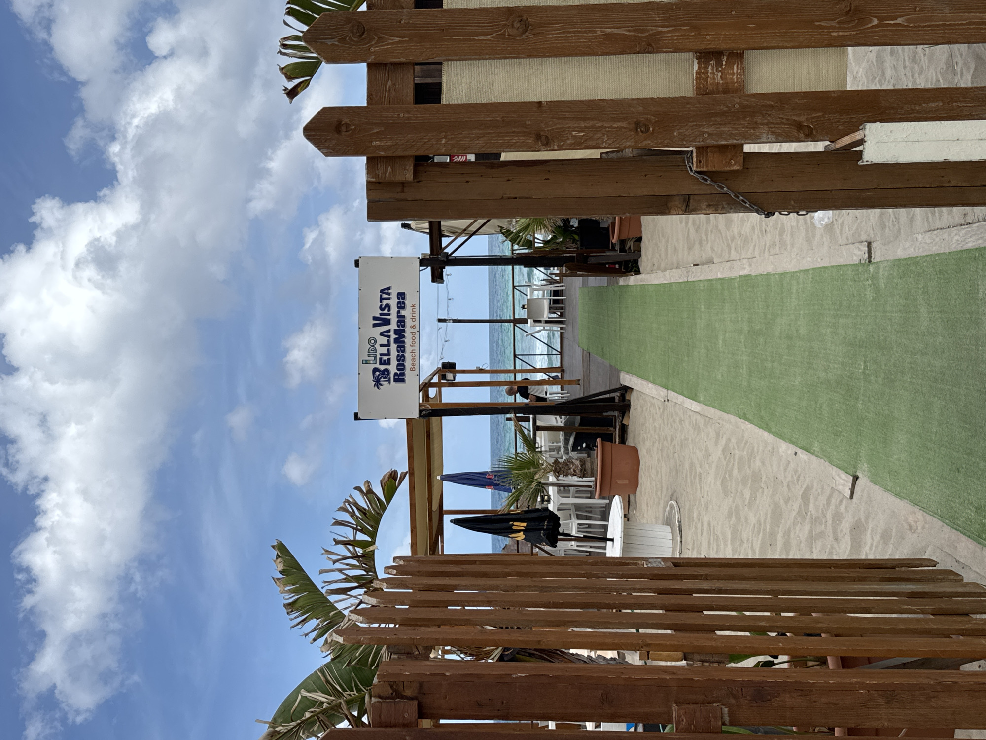 An exterior shot captures a beachside establishment in Isola delle Femmine, Italy. In the foreground, a wooden gate made of vertical planks stands open, revealing a sandy path lined with a green artificial turf runner. To the right of the path, a section of the wooden fence is covered with a beige canvas material.
Beyond the gate, tables and chairs are arranged on a raised wooden deck, some covered by a striped awning. Umbrellas are visible, including a black one with yellow branding and two blue ones. A sign overhead reads "LIDO BELLA VISTA RosaMarea" with the text "Beach food & drink" below. The sea stretches out in the background under a partly cloudy sky.
In the middle ground, a person is seated at one of the tables. The environment suggests a relaxed, outdoor dining or lounge area. The overall scene depicts a functional beachside venue ready to serve patrons.